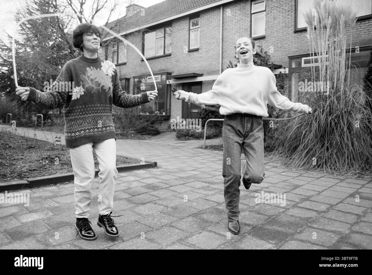 Familie Thijssen Springseil Badhoevedorp Badhoevedorp, Whizgle News, Dutch Desk, Niederlande, 1950 - 2000 am 23-01-1991. Diese Themen sind in der Abbildung dargestellt. In einer malerischen Umgebung im Freien können sich zwei Personen freudig an Seilspringen beteiligen und eine Atmosphäre voller Überschwänglichkeit und Verspieltheit schaffen. Die Szene wird von einer Kulisse aus bescheidenen Backsteinhäusern eingerahmt, die durch große Fenster gekennzeichnet sind, die eine gemütliche Wohnumgebung suggerieren. Der Boden ist mit quadratischen Steinen gepflastert, die in einem ordentlichen Muster angeordnet sind und zu gepflegten Gartenflächen führen, die den Weg flankieren. Hohe Gräser und niedrige Stockfoto