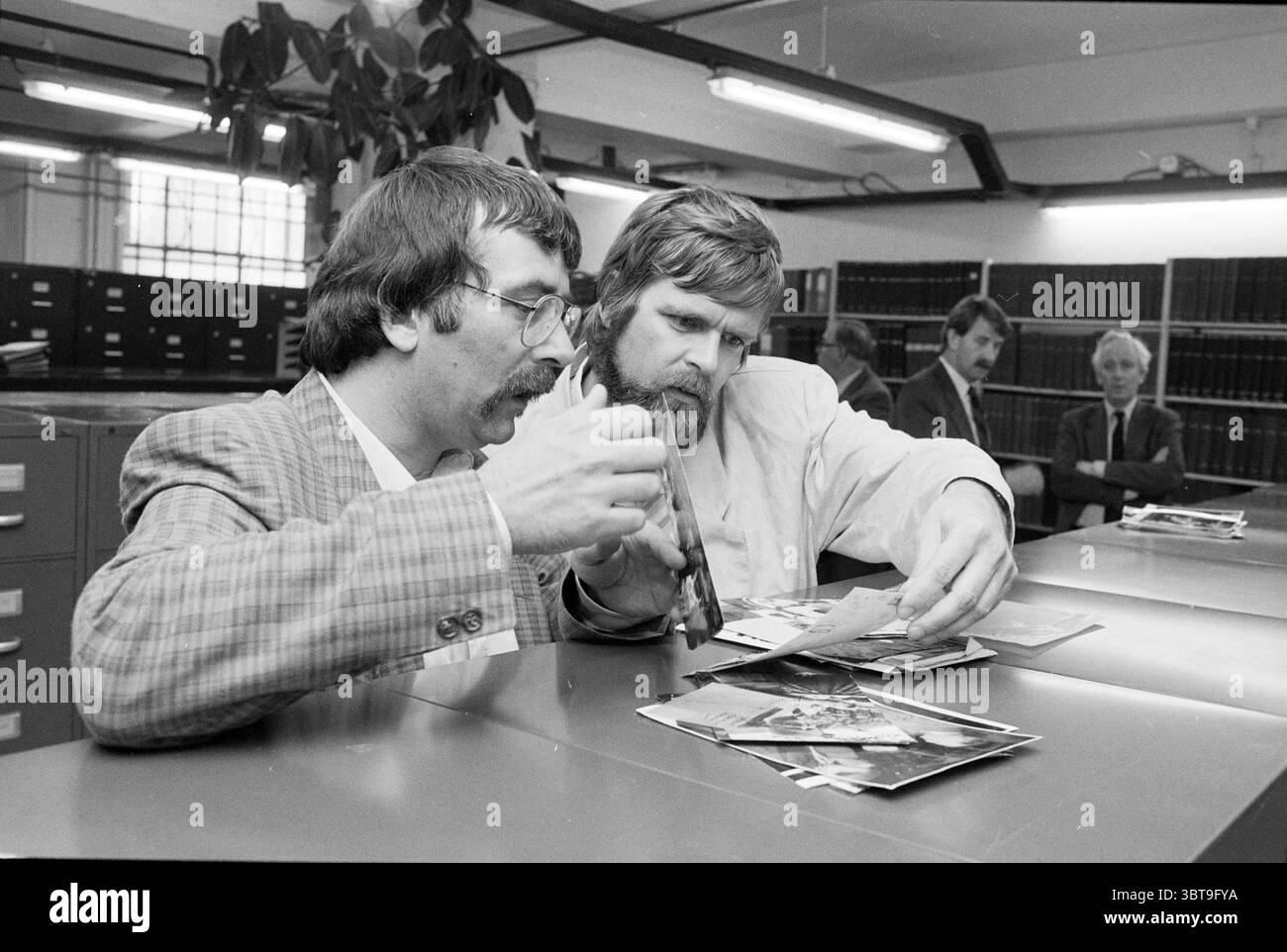 B&W Haarlem at Spaarnestad Archives Haarlem Municipal Council, Whizgle News, Dutch Desk, Niederlande, 1950 - 2000 am 26-05-1987. Dies sind die Elemente im Bild. In einem gut beleuchteten Innenraum führen zwei Männer eine konzentrierte Diskussion über eine Sammlung von Fotografien, die auf einer Ebenen Fläche angeordnet sind. Der Mann auf der linken Seite, gekleidet in einem hellgrauen Karomuster, trägt einen gepflegten Schnurrbart und wirkt besonders animiert, als er mit einer Schere in einer Hand Gesten macht, die einen Punkt betonen. Sein Ausdruck deutet auf Neugier und Interesse hin, während seine andere Hand auf die Fotografien zeigt Stockfoto