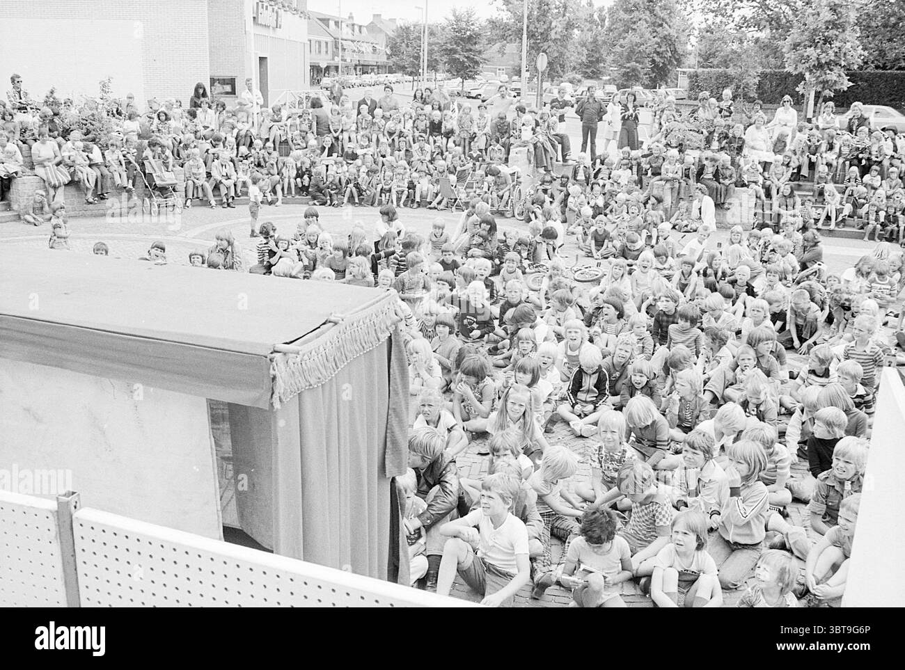 Puppentheater Nielenplein H'kerk Puppentheater Puppentheater Heemskerk Burgemeester Nielenplein, Whizgle News, Dutch Desk, Niederlande, 1950 - 2000 am 13-07-1977. Dies sind die Elemente im Bild. Die Szene ist gefüllt mit einer großen Menge von Kindern, die alle zusammen mit einem spürbaren Gefühl von Aufregung und Neugier versammelt sind. Sie sitzen eng auf dem Boden und schaffen ein Meer jugendlicher Gesichter und abwechslungsreicher Frisuren, die von kurzen Frisuren bis hin zu langen, fließenden Styles reichen. Die Kinderbekleidung zeigt eine Mischung aus Mustern und Farben, viele tragen farbenfrohe Hemden Stockfoto