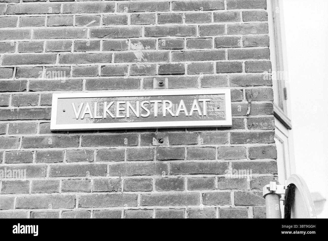 Zwei Valkestraat-Schilder Haarlem Valkestraat Niederlande, Whizgle News, Dutch Desk, Niederlande, 1950 - 2000 am 08-06-1992. Dies sind die Elemente im Bild. Die Szene zeigt eine strukturierte Wand aus kleinen, rechteckigen Ziegeln, die in einem klassischen Muster angeordnet sind. Die Ziegelsteine sind hauptsächlich in gedämpftem Grau gehalten und weisen Farbvariationen auf, die auf Alter und Verschleiß hinweisen. Die Oberfläche zeigt subtile Anzeichen von Verwitterung, mit einigen Bereichen, in denen der Mörtel abgesplitzt oder verfärbt ist. Der Schwerpunkt der Komposition ist ein rechteckiges Metallplakat, das an der Wand angebracht ist. Das Plakat hat eine polierte silberne Oberfläche. Stockfoto