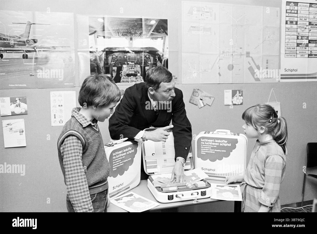 Präsentation des Luftfahrtlehrkoffer Schiphol Schiphol, Whizgle News, Dutch Desk, Niederlande, 1950 - 2000 am 23-04-1980. Diese Themen werden in der Abbildung angezeigt. In einer Umgebung, die an ein Klassenzimmer oder einen informativen Ausstellungsbereich erinnert, steht ein Mann mittleren Alters in dunkler Uniform in der Mitte, der zwei Kinder begeistert. Der Ausdruck des Mannes vermittelt ein Gefühl von Autorität, gemischt mit Wärme, während er auf einen großen Koffer zeigt, der vor ihm offen gelegt wurde. Dieser Fall ist mit verschiedenen Artikeln und Schulungsmaterialien gefüllt, die eine Präsentation oder Demonstration zu einem verwandten Thema vorschlagen Stockfoto