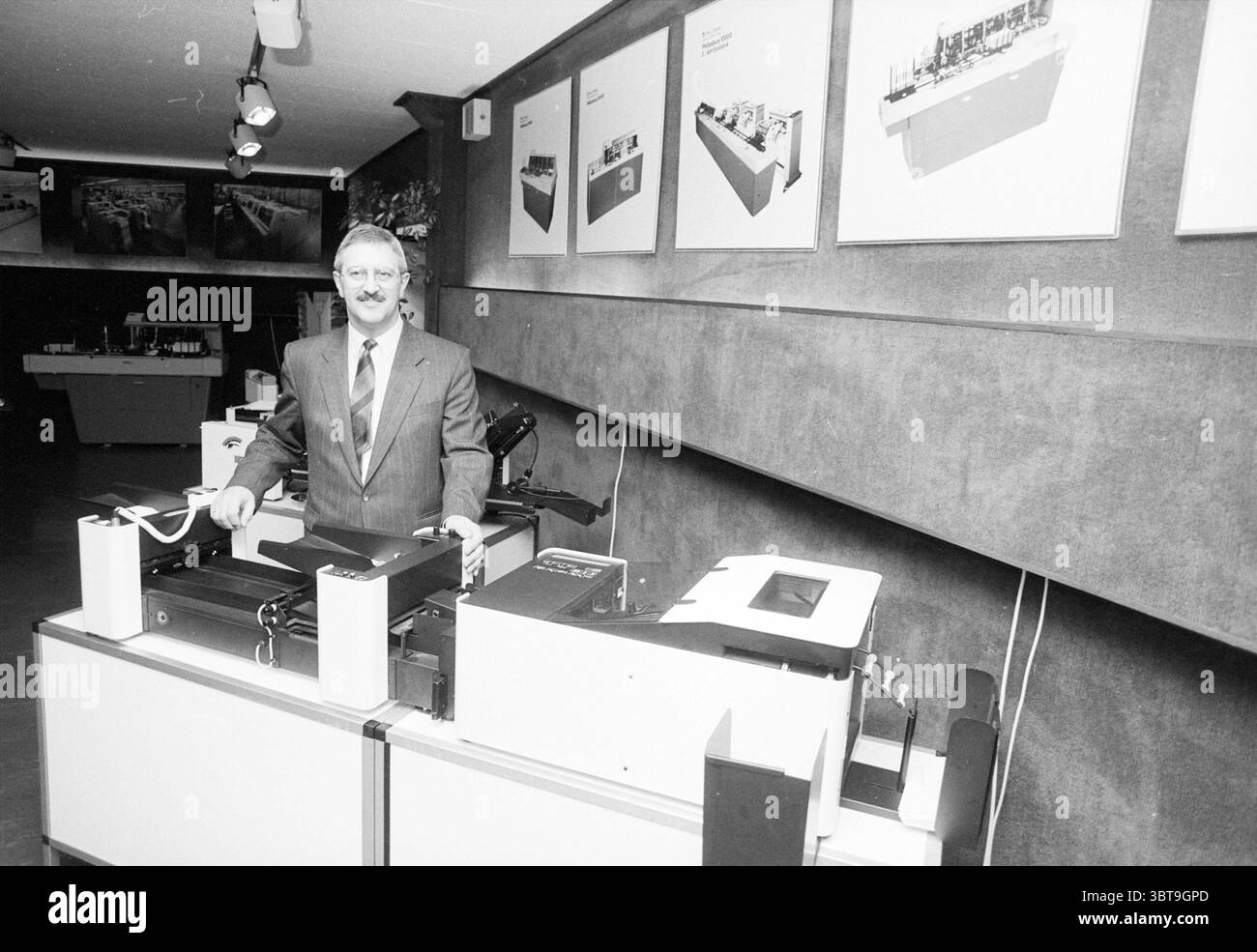 Herr Korsting Pretech Zwanenburg Persons Companies Zwanenburg, Whizgle News, Dutch Desk, Niederlande, 1950 - 2000 am 20-01-1989. Diese Themen sind in der Abbildung dargestellt. In einem geschäftlich anmutenden Innenraum steht ein gut gekleideter Mann neben einer großen, anspruchsvollen Maschine, die wahrscheinlich mit Technologie oder Druck zu tun hat. Er trägt einen maßgeschneiderten Anzug, der Professionalität vermittelt und ein subtiles Lächeln hat, was auf Selbstvertrauen hindeutet. Die Umgebung ist mit einer gedämpften Palette von Grautönen gefüllt, die durch die helle, gleichmäßige Beleuchtung über dem Kopf hervorgehoben wird, wodurch eine saubere und organisierte Atmosphäre entsteht. Hinter ihm, eine Reihe von gerahmten Illustr Stockfoto