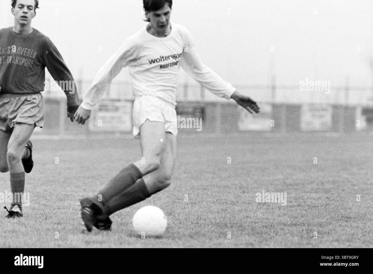 Fußballspiel Hoofddorp, Whizgle News, Dutch Desk, Niederlande, 1950 - 2000 am 02-1987. Diese Themen werden in der Abbildung angezeigt. Die Szene fängt ein Fußballspiel im Freien ein, das durch einen bewölkten, bewölkten Himmel gekennzeichnet ist, der ein gedämpftes Licht über das Feld wirft. Das Gras ist feucht und wirkt trotz des düsteren Wetters satt und lebhaft. Im Vordergrund ist ein Spieler in Bewegung und streckt sein Bein anmutig aus, während er mit einem weißen Fußball in Kontakt kommt. Er trägt ein helles Trikot mit langen Ärmeln, ein Logo, das nicht zu erkennen ist, und er trägt dunkle Shorts. Seine ausdrucksstarke Haltung suggeriert Stockfoto