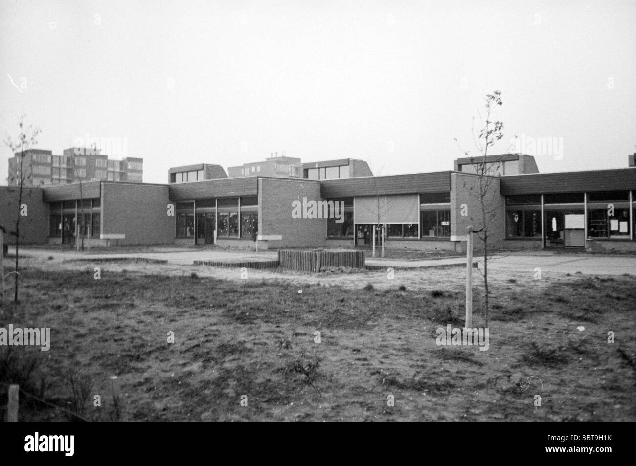 Außenkindergarten de Vrijbuiters Hoofddorp Vorschulkindgärten, Whizgle News, Dutch Desk, Niederlande, 1950 - 2000 am 21-12-1977. Das Bild enthält diese Themen. Die Szene zeigt ein Schulgebäude, das sich durch seine moderne Architektur auszeichnet und eine flache Struktur mit mehreren rechteckigen Abschnitten aufweist. Die Außenwände bestehen aus gedämpften rötlich-braunen Ziegeln, im Kontrast stehen große, horizontale Fenster, die auf natürliches Licht hinweisen. Die flachen Dachlinien und kantigen Formen verleihen dem Design ein modernes Flair. Im Vordergrund breitet sich ein Stück karger Erde aus, Dott Stockfoto