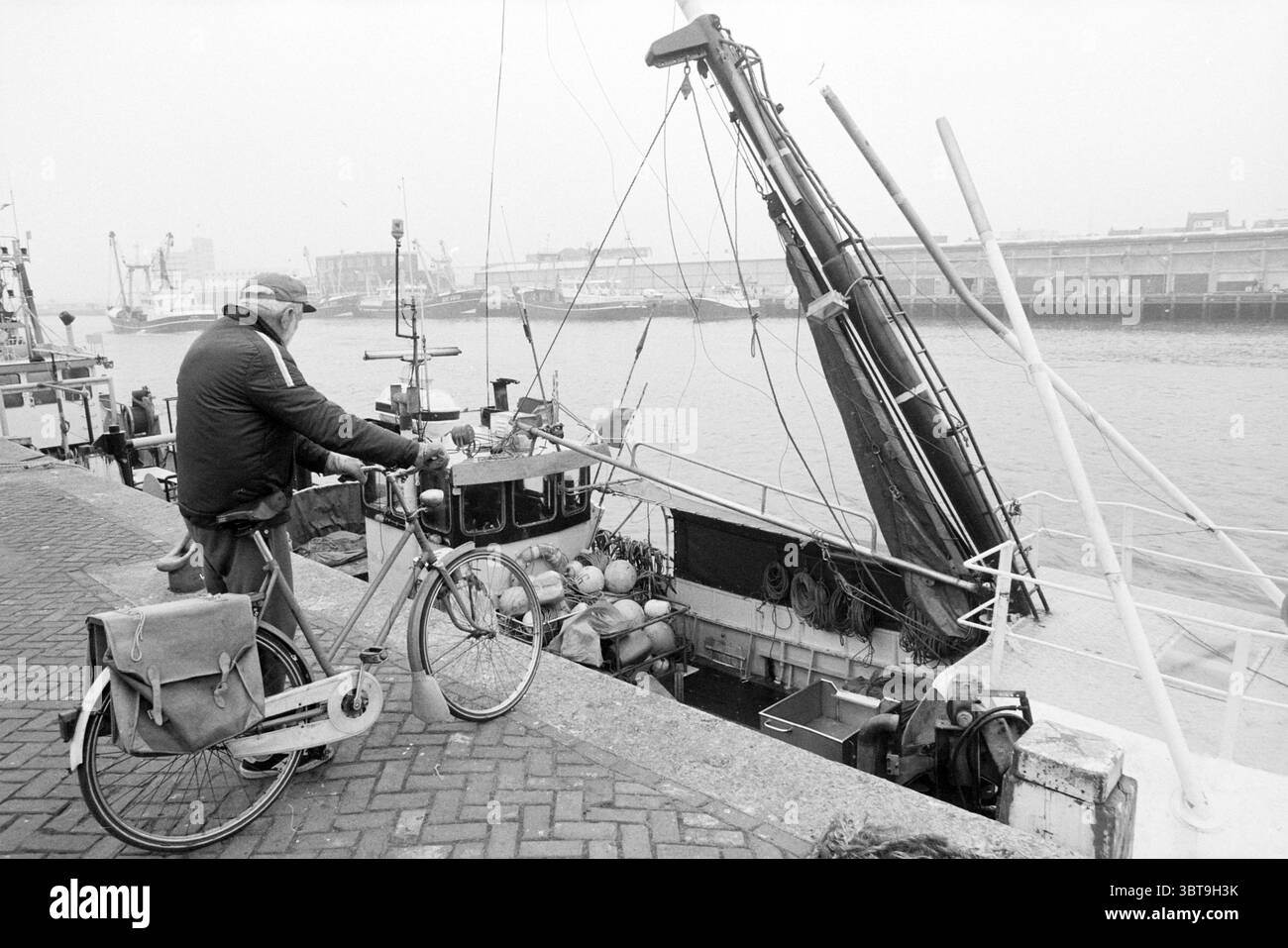 Cutter IJM 39 mit gebrochenem Mast IJmuiden Niederlande, Whizgle News, Dutch Desk, Niederlande, 1950 - 2000 am 08-11-1994. Die Abbildung zeigt diese Themen. Die Szene zeigt einen Küstenhafen, umgeben von einer gedämpften, nebeligen Atmosphäre, die einen Hauch von Ruhe und Nostalgie verleiht. Die Farbpalette ist überwiegend Graustufen, mit Anklängen von weichem weiß und tiefen Schatten, die die ruhige, aber leicht melancholische Stimmung verstärken. Im Vordergrund steht ein älteres Individuum neben einem Fahrrad, das auf einem strukturierten Steinsteg geparkt ist. Der Mann trägt eine dunkle Jacke, mit einer tief gezogenen Mütze, die seine teilweise verdeckt Stockfoto