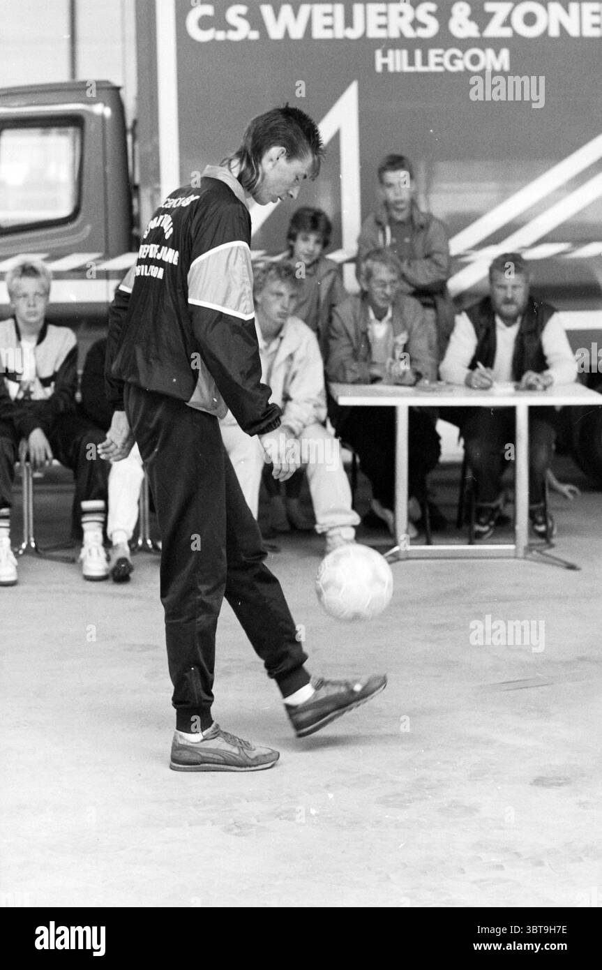 Gert Jan Oudshoorn hält den Weltrekordball aufrecht Hillegom Record Improves and Improvement Hillegom, Whizgle News, Dutch Desk, Niederlande, 1950 - 2000 am 25.07.1987. Diese Themen werden in der Abbildung angezeigt. In einem geräumigen Innenbereich sitzt eine Gruppe von Einzelpersonen an einem langen Tisch, deren Aufmerksamkeit sich intensiv auf eine Person konzentriert, die im Vordergrund steht. Diese zentrale Figur demonstriert eindrucksvolle Ballkontrolle und balanciert lässig einen Fußball auf einem Fuß. Ihre sportliche Kleidung besteht aus einer dunklen Jacke mit kontrastierenden weißen Abschnitten und einer taillierten Hose, die auf eine Hingabe an den Sportler hinweist Stockfoto