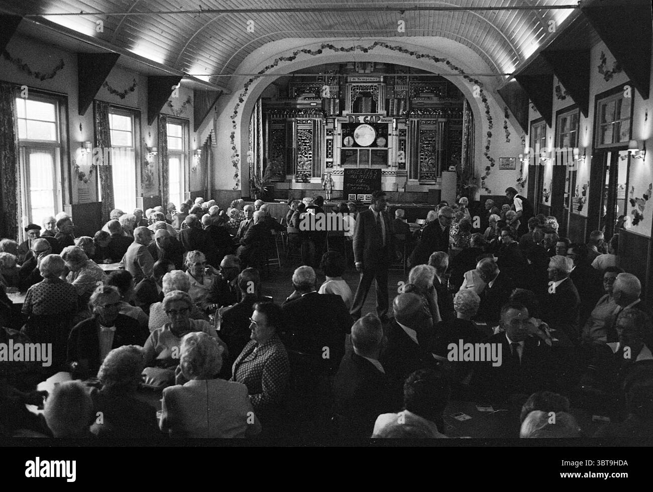 Bankettsaal mit Orgelsäulen, Whizgle News, Dutch Desk, Niederlande, 1950 - 2000 am 07-06-1972. Die Abbildung zeigt diese Themen. Die Szene zeigt einen belebten Saal mit einer großen Versammlung von Menschen, überwiegend älteren Erwachsenen, die eng um Tische sitzen. Der Raum zeichnet sich durch eine hohe Bogendecke aus, die den Blick nach oben zieht, mit Dekorationen und Mustern, die ein Gefühl von Tradition und Geschichte vermitteln. Die Farbpalette ist weitgehend monochromatisch und orientiert sich an Grautönen, was eine zeitlose, vielleicht nostalgische Stimmung vermittelt. Licht strömt von großen Fenstern, die den einrahmen Stockfoto