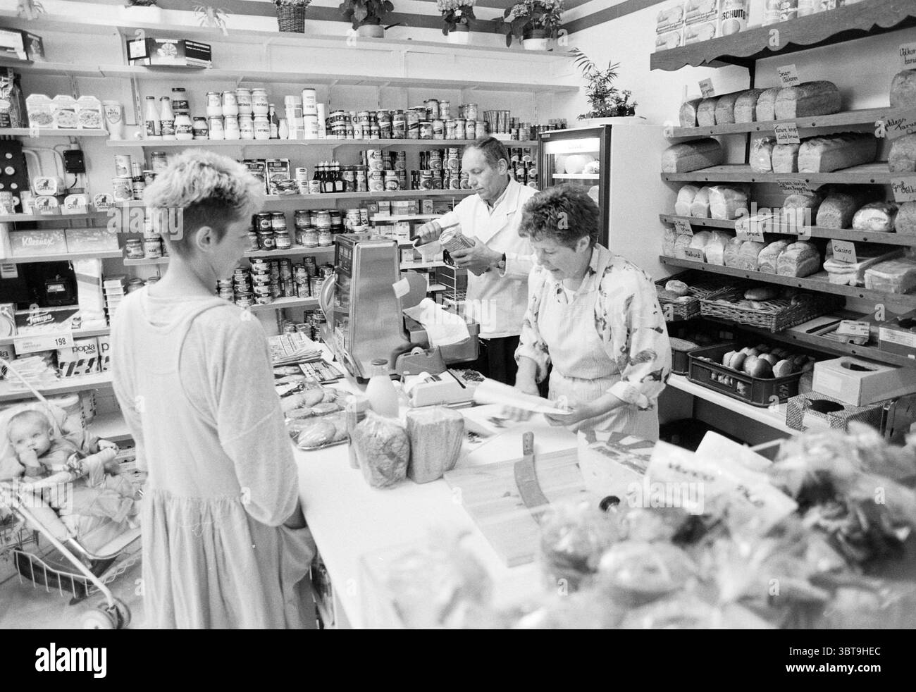 Der Laden in der Nachbarschaft war fantastisch. Noom Neighborhoods Community Centers örtliche Geschäfte Trainingszentren, Whizgle News, Dutch Desk, Niederlande, 1950 - 2000 auf 30-04-1990. Dies sind die Elemente im Bild. In einem hübschen Lebensmittelgeschäft verströmt die Inneneinrichtung einen wohnlichen Charme mit Regalen voller Gläser, Dosen und verschiedenen verpackten Waren. Die Wände sind mit Holzregalen gesäumt, in denen eine Auswahl an Lebensmitteln ausgestellt ist. In der Mitte befindet sich eine Theke, in der eine Frau mit lockigem Haar, die eine Schürze trägt, mit dem Auschecken eines Kunden beschäftigt ist. Sie hat einen fokussierten Ausdruck, da sie Gegenstände auf einem alten fas abwägt Stockfoto