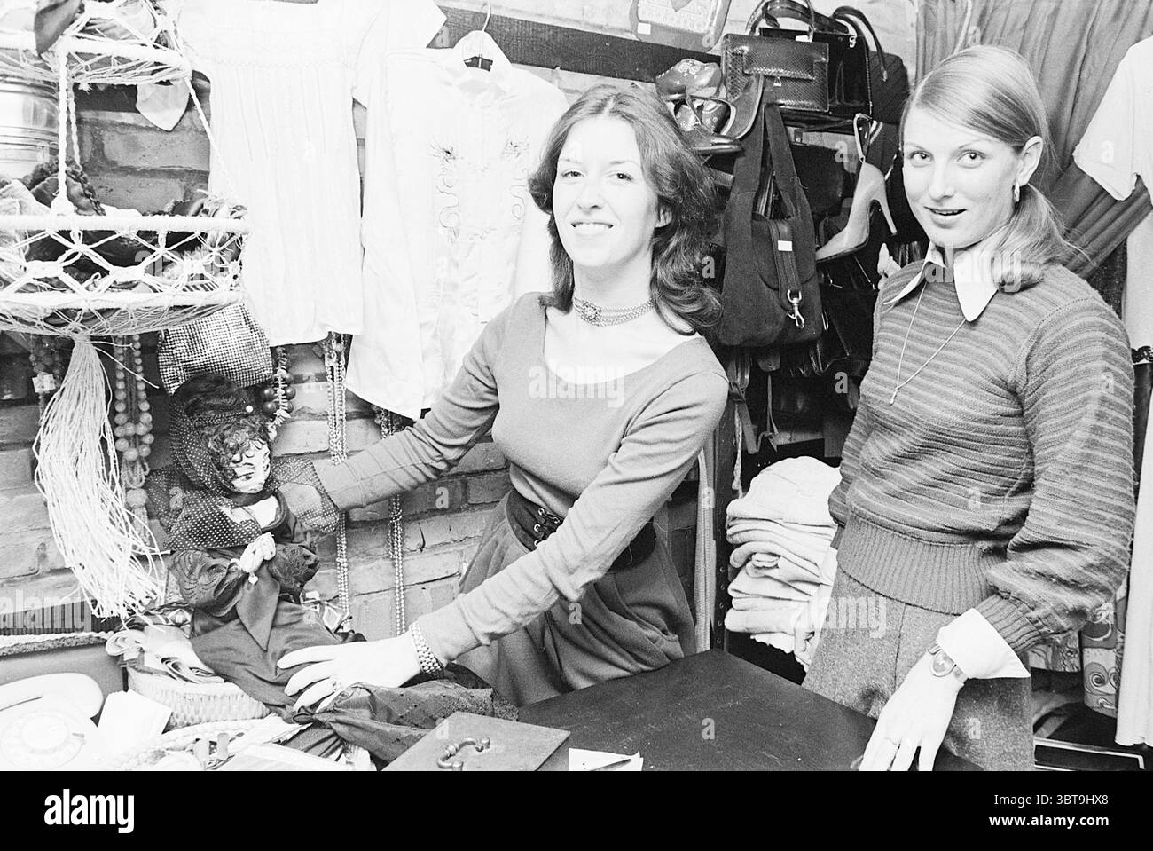 Second-Hand-Bekleidungsgeschäft Beverwijk Clothing, Whizgle News, Dutch Desk, Niederlande, 1950 - 2000 am 08-03-1977. Dies sind die Elemente im Bild. In der Szene befinden sich zwei Frauen in einer malerischen, Vintage-Umgebung, die mit verschiedenen Objekten gefüllt ist. Die erste Frau links steht mit einem freundlichen Lächeln und zeigt eine kleine Puppe in dunkler Kleidung. Ihr langes, gewelltes Haar umrahmt ihr Gesicht und sie trägt ein langärmeliges Kleid, das sanft drapiert ist und eine gedämpfte, warme Farbe hat. Die zweite Frau rechts steht leicht abgewinkelt und gibt einen spielerischen Ausdruck. Ihr Haar ist glatt, Stockfoto