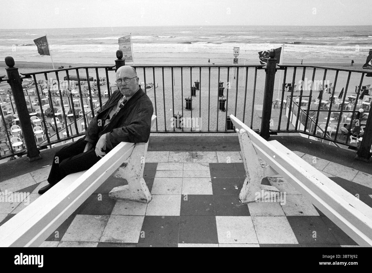 Herr Paap Tourismus Zandvoort [Tourist] Zandvoort, Whizgle News, Dutch Desk, Niederlande, 1950 - 2000 am 22-06-1995. Das Bild enthält diese Themen. Die Szene zeigt einen düsteren Blick auf den Strand, mit einer Einzelfigur auf einer verwitterten Bank, die weiß gestrichen wurde, aber jetzt etwas verblasst und abgesplittert ist. Der ältere Mann, gekleidet in dunkler Jacke und Brille, sitzt besinnlich und blickt auf den weiten Ozean in der Ferne. Seine Anwesenheit schafft ein starkes Gefühl der Einsamkeit. Der Hintergrund zeigt ein Sandstrand, gespickt mit verschiedenen Strandhütten und Sonnenschirmen, Sugges Stockfoto