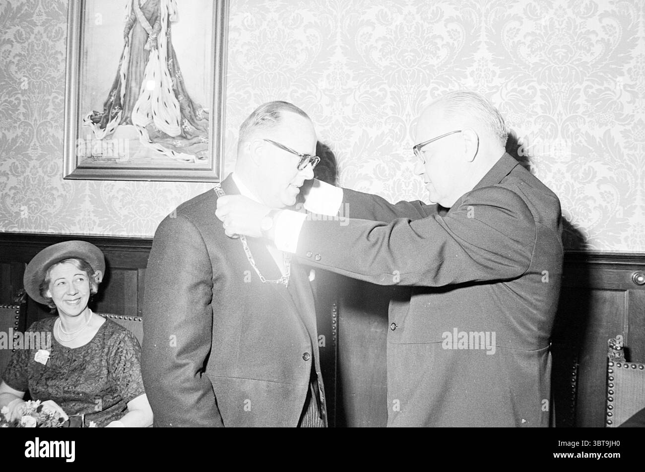 Installationsbürgermeister Niekerk in Hillegom. Installation, Whizgle News, Dutch Desk, Niederlande, 1950 - 2000 am 06-06-1964. Das Bild enthält diese Themen. In einem reich dekorierten Raum, der mit kunstvollen Tapeten mit Blumenmuster geschmückt ist, findet eine formelle Veranstaltung statt. Die Umgebung strahlt durch ihre kunstvollen Details und die gedämpfte, aber warme Beleuchtung einen Hauch von Eleganz aus. Zwei Männer sind im Zentrum des Geschehens. Der eine trägt einen dunklen Anzug und steht stolz wie der andere, auch in formeller Kleidung, legt ihm eine zeremonielle Kette um den Hals, was auf eine Verleihung oder Anerkennung hindeutet. Das Gentleman-Wesen Stockfoto