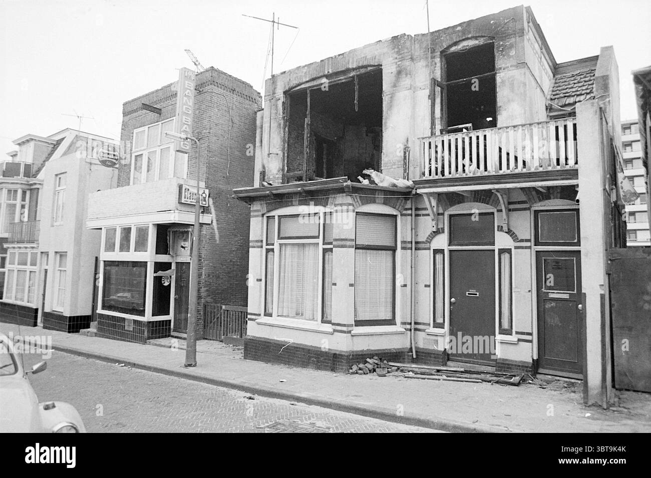 Ausgebranntes Gebäude neben der Bar Bodega 'The Bang Bang' Zandvoort Stationsstraat, Whizgle News, Dutch Desk, Niederlande, 1950 - 2000 am 04-1974. Dies sind die Elemente im Bild. Die Szene präsentiert eine raue urbane Umgebung, die durch zwei benachbarte Gebäude in einer ruhigen Straße gekennzeichnet ist. Auf der linken Seite hat ein flaches Dach ein einfacheres Design mit großen Fenstern mit dekorativen Zierleisten. Die Fassade ist mit einer Mischung aus weißer und heller Farbe geschmückt, die jetzt etwas verwittert ist. Daneben fällt ein aufwändigeres Gebäude auf. Es hat eine halb ruinierte obere Ebene mit exponiertem bri Stockfoto