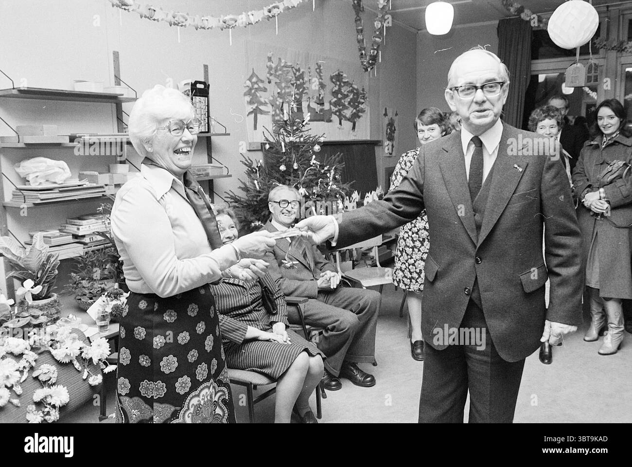 Kinder schenken älteren Menschen Weihnachtsstücke. Weihnachten und Weihnachtsbäume Kinder Kinder Kinder Kinderheime Kinder Party Kinder, Whizgle News, Dutch Desk, Niederlande, 1950 - 2000 am 15-12-1976. Dies sind die Elemente im Bild. Die Szene fängt ein festliches Treffen ein, wahrscheinlich während einer Feier oder Feiertagsfeier. Der Fokus liegt auf einem älteren Paar, das an vorderster Front steht und sich in einer freudigen Interaktion engagiert. Die Frau, gekleidet in einem gemusterten Rock und einem hellen Top, lächelt weit und strahlt Wärme und Glück aus. Sie hält ihre Hand aus und scheint dem Mann neben ihm etwas zu präsentieren Stockfoto
