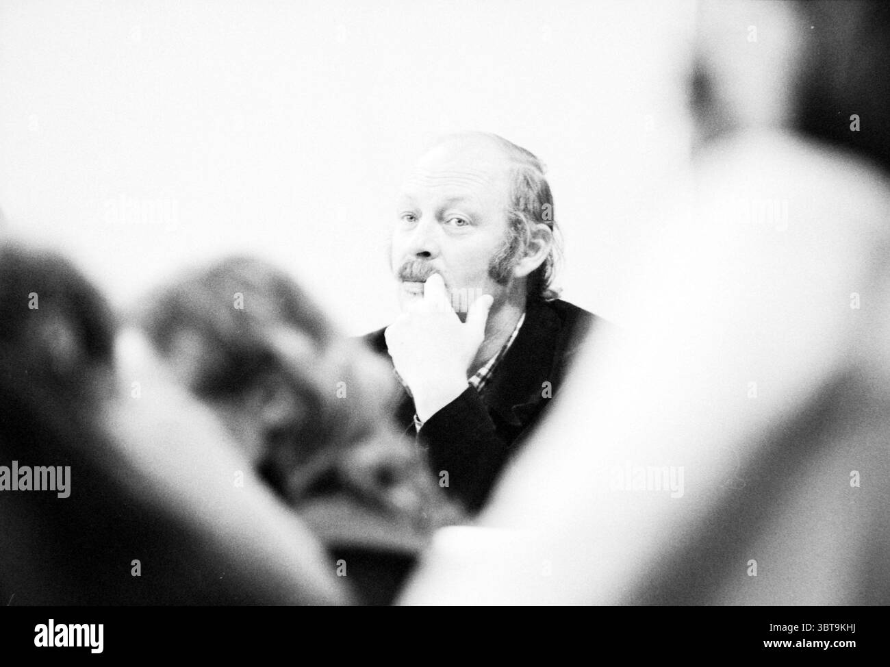 Budget Haarlem Haarlem Gemeinderat Haarlem the Netherlands, Whizgle News, Dutch Desk, Niederlande, 1950 - 2000 am 05-11-1980. Dies sind die Elemente im Bild. Die Szene ist eine monochromatische Darstellung, die Grautöne betont, die eine nostalgische oder zeitlose Stimmung erzeugen. Im Zentrum der Komposition steht ein älterer Mann, der sich durch seinen nachdenklichen Ausdruck und seine teilweise gefurchten Augenbrauen auszeichnet. Er hat eine dünnere Mähne, die auf eine abfallende Haarlinie hindeutet, und einen ordentlich getrimmten Schnurrbart. Seine Hand liegt nachdenklich auf seinem Kinn, was auf tiefe Besinnung hindeutet. Um ihn herum sind verschwommene Figuren Stockfoto