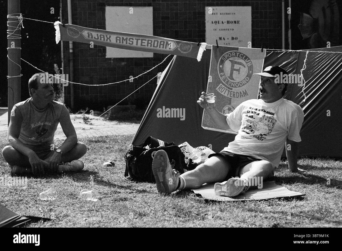 Sonstiges Beach 6 - Day, Whizgle News, Dutch Desk, Niederlande, 1950 - 2000 am 28-07-1992. Diese Themen sind in der Abbildung dargestellt. In der Umgebung sitzen zwei Männer in einem grasbewachsenen Bereich, was eine ungezwungene und entspannte Atmosphäre verkörpert. Das Sonnenlicht taucht die Szene in ein warmes Licht und sorgt für eine entspannte Stimmung. Ein Mann, gekleidet in einem weißen T-Shirt und Shorts, lehnt sich bequem und schlürft aus einem klaren Glas, während sein Ausdruck ein Gefühl von Freizeit vermittelt. Seine Kleidung deutet auf einen sportlichen Lebensstil hin, und er scheint in Gesprächen verwickelt zu sein. Der andere Mann, der ein dunkleres Hemd und Shorts trägt, sitzt im Kreuz ne Stockfoto