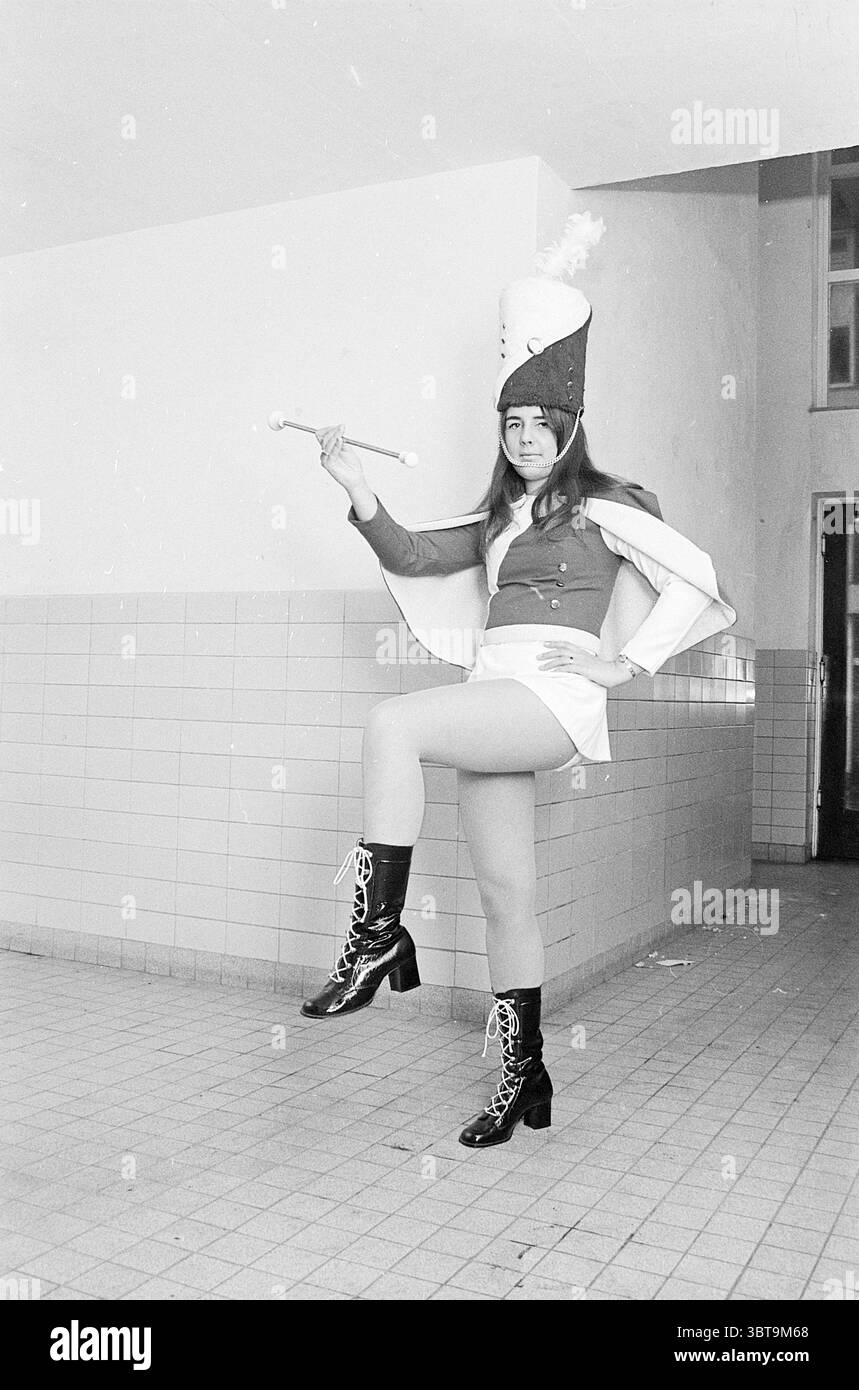 Majorette Music, Whizgle News, Dutch Desk, Niederlande, 1950 - 2000 auf 31-10-1970. Diese Themen sind in der Abbildung dargestellt. Ein junger Mensch steht selbstbewusst in einem Innenraum, der von starken, neutralen Wänden und Fliesenböden geprägt ist. Sie tragen eine auffällige Uniform, die an ein Marschband oder ein Drillteam erinnert. Sie verfügt über eine eng anliegende Jacke mit einer auffälligen Farbe, die durch eine weiße Schärpe ergänzt wird, die über eine Schulter fällt. Das Outfit ist mit einem hohen, zeremoniellen Hut mit einer Feder versehen, die ein zeremonielles Flair verleiht. Die Person schlägt eine Pose mit einem erhobenen Bein und zeigt t Stockfoto
