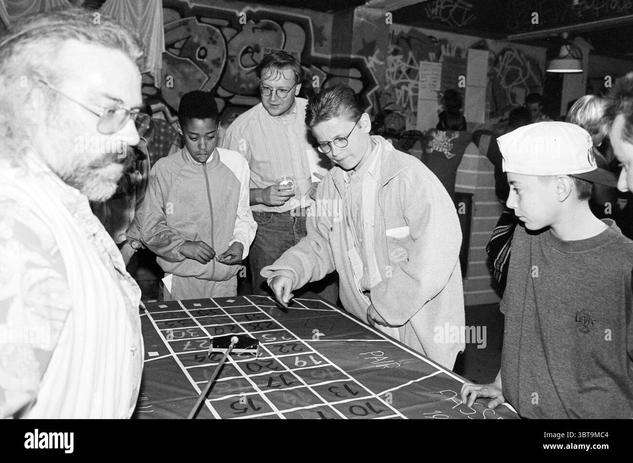 Spielsucht Themenabend IJmuiden IJmuiden the Netherlands, Whizgle News, Dutch Desk, Niederlande, 1950 - 2000 auf 30-04-1995. Dies sind die Themen im Bild. Die Szene ist voller Energie und spielt in einem lebendigen Innenraum, der von einer eklektischen Mischung aus Persönlichkeiten und Aktivitäten geprägt ist. Die Kulisse zeigt ein visuell chaotisches Arrangement von Graffiti, das helle Farben und dynamische Designs zeigt, die für eine urbane, kreative Umgebung sprechen. Im Vordergrund versammeln sich mehrere Personen um einen großen Tisch mit einem Spiellayout, der scheinbar in ein Spiel vertieft ist, das Chan beinhaltet Stockfoto