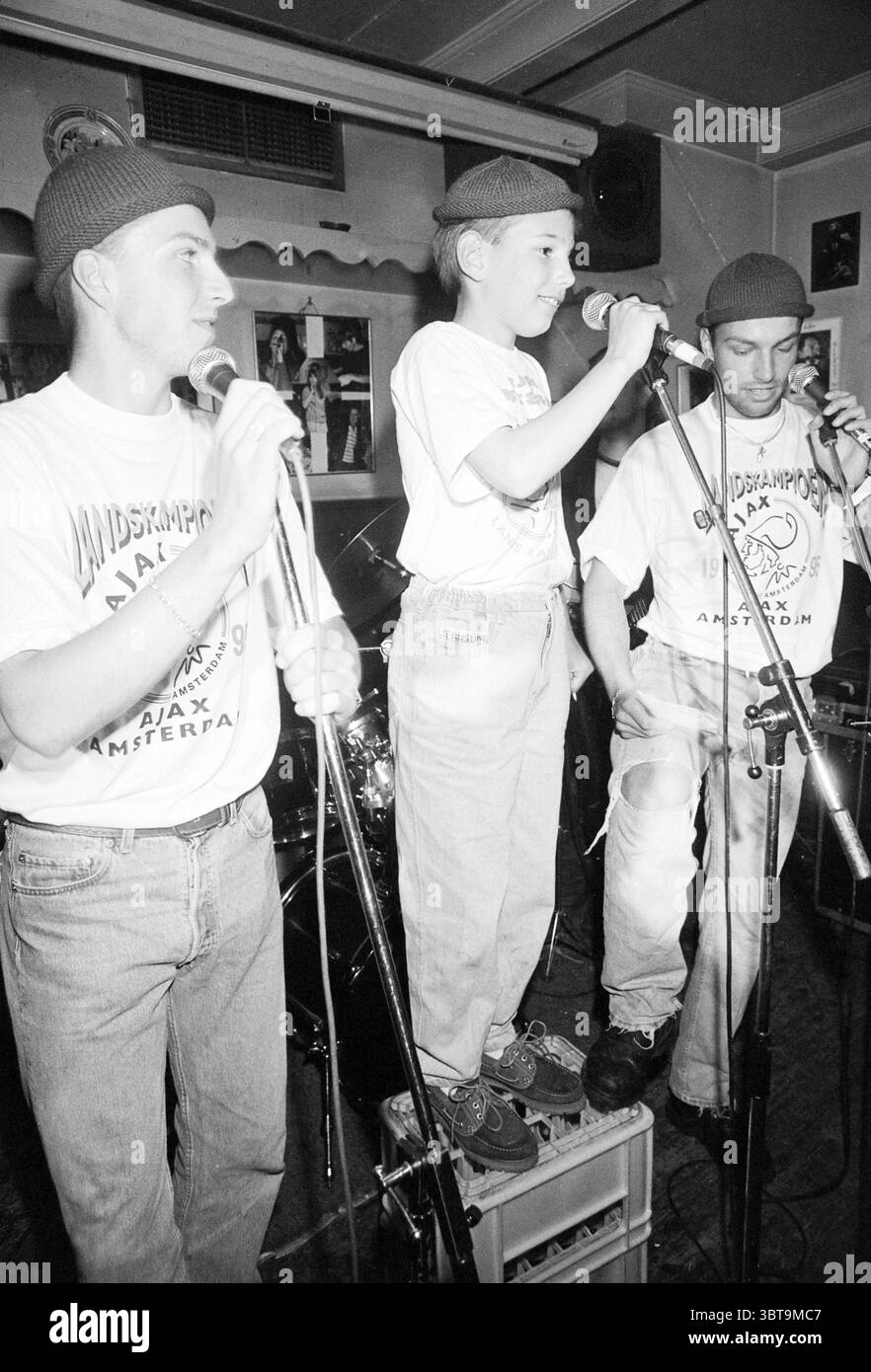 Themenabend „You CAN do this on Stage Today“ in Taverna Imperial Korte Veerstraat 3 Haarlem Korte Veerstraat Nederland, Whizgle News, Dutch Desk, Niederlande, 1950 - 2000 am 12.05.1995. Das Bild enthält diese Themen. In einem lebendigen Innenbereich stehen drei Personen an prominenter Stelle, die an einer Vorstellung beteiligt sind. Die Szene ist thematisch lebendig und dynamisch und schafft eine Atmosphäre von Enthusiasmus und Energie. Die beiden Erwachsenen auf beiden Seiten tragen eine ähnliche Kleidung: Lockere, leicht verblasste Jeans und weiße T-Shirts mit auffälligen Grafikdesigns. Beide Männer tragen passende dunkle Kappen Stockfoto