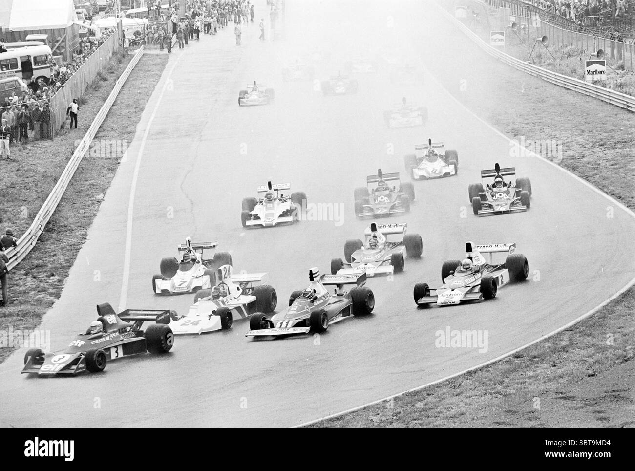 Grand Prix in Zandvoort Grand Prix, Whizgle News, Dutch Desk, Niederlande, 1950 - 2000 am 22-06-1975. Das Bild enthält diese Themen. Die Szene fängt einen dynamischen Moment eines klassischen Motorrennens ein, bei dem mehrere Rennwagen mit hoher Geschwindigkeit auf einer kurvenreichen Strecke fahren. Die Fahrzeuge sind schlank und niedrig und bieten eine Farbmischung wie tiefes Rot, Hellblau und klassisches weiß, jeweils mit markanten Rennnummern und Sponsorenlogos. Die Kulisse ist eine Rennstrecke, die mit hohem Gras und Barrieren gesäumt ist, die eine Kulisse der Spannung schafft. Ein weicher Dunst umhüllt die Szene, wahrscheinlich vom Tire smo Stockfoto