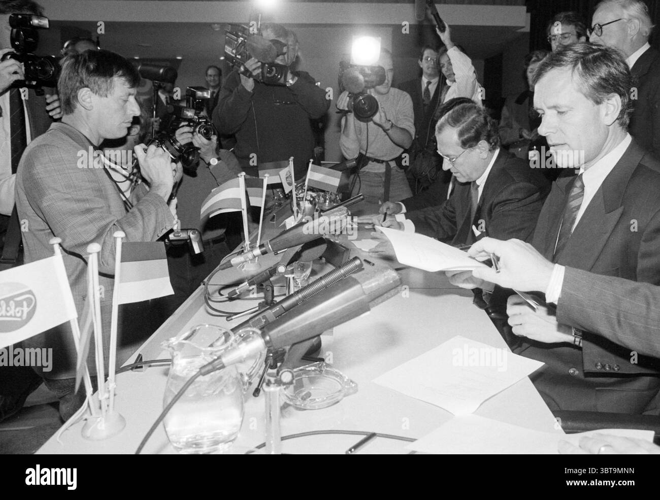 Vertrag Fokker & Dasa, Whizgle News, Dutch Desk, Niederlande, 1950 - 2000 am 29-10-1992. Dies sind die Elemente im Bild. In einer formellen Umgebung befindet sich in der Mitte ein runder Tisch, an dem zwei Männer sitzen. Die Atmosphäre vermittelt ein Gefühl von Wichtigkeit und Spannung, da mehrere Kameras und Journalisten die Szene umgeben und jeden Moment festhalten. Die Männer scheinen konzentriert zu sein, einer von ihnen ist dabei, ein Dokument zu unterzeichnen, mit Papieren vor ihm verteilt. Die Einzelpersonen sind in formeller Kleidung gekleidet: Anzüge und Krawatten, was auf eine Verhandlung mit hohem Stellenwert hindeutet. Die Beleuchtung ist hell und unterstreicht den Stockfoto