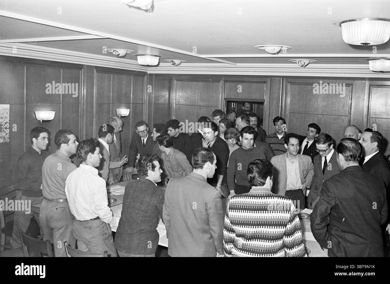 Meeting of Spanish Guest Workers and Other Men., Whizgle News, Dutch Desk, Niederlande, 1950 - 2000. Diese Themen sind in der Abbildung dargestellt. Die Szene entfaltet sich in einem gut beleuchteten Raum, der durch eine warme Holztäfelung gekennzeichnet ist, die ein Gefühl von Intimität verleiht. Eine Gruppe von Einzelpersonen trifft sich eng zusammen, wobei ihr Fokus auf einen zentralen Tisch gerichtet ist, an dem Diskussionen oder Aktivitäten stattfinden. Es gibt etwa zwei Dutzend Menschen, einige stehen und andere sitzen, die alle in einer Reihe von Stilen gekleidet sind, die typisch für diese Zeit sind, überwiegend in dunklen und gedämpften Farben. Die Einzelpersonen exhi Stockfoto