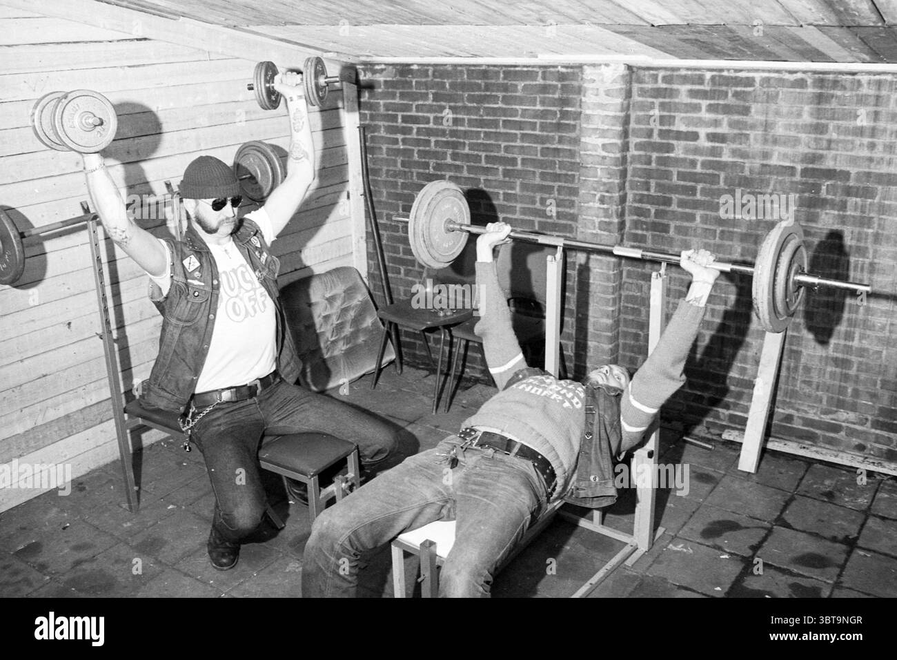 Hells Angels in der Turnhalle des Jugendzentrums Het Anker Parklaan 45 Haarlem. Haarlem Parklaan the Netherlands, Whizgle News, Dutch Desk, Niederlande, 1950 - 2000 am 20-06-1980. Das Bild enthält diese Themen. In einem schwach beleuchteten Raum nehmen zwei Personen an einem Gewichtheben Teil. Eine Person sitzt auf einem Stuhl, hebt eine Langhantel mit einem fokussierten Ausdruck über den Kopf und zeigt, wie die Armmuskulatur gegen das Gewicht anstrengt. Sie tragen eine dunkle Sonnenbrille und eine ärmellose Jeansweste mit Patches, die auf einen robusten, rebellischen Stil hinweisen. Die zweite Person liegt auf einer Bank und bereitet sich darauf vor Stockfoto