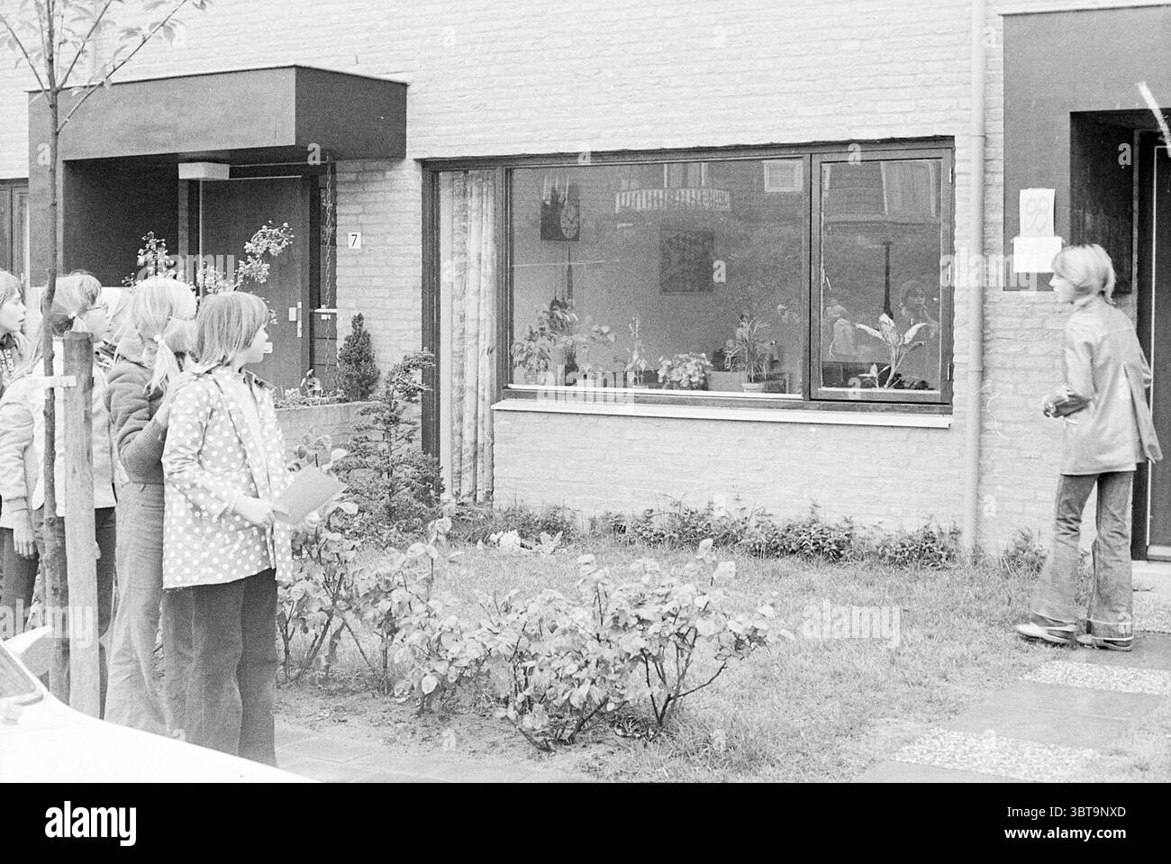Festlichkeiten Noordwijkerhout Queen's Day, Whizgle News, Dutch Desk, Niederlande, 1950 - 2000 auf 30-04-1974. Dies sind die Themen im Bild. In der Szene steht eine Gruppe von mehreren Personen in einer Reihe entlang eines Gehwegs, gegenüber einem Gebäude mit großen, rechteckigen Fenstern. Die einzelnen Personen scheinen an einer gemeinsamen Aktivität beteiligt zu sein und zeigen Ausdruck von Neugier und Vorfreude. Die Bekleidungsstile lassen eine lässige, möglicherweise Retro-Ästhetik vermuten, wobei eine Person ein gepunktetes Hemd trägt, was der gesamten Szene ein verspieltes Element verleiht. Das Gebäude selbst hat ein modernes, minimalistisches Design Stockfoto