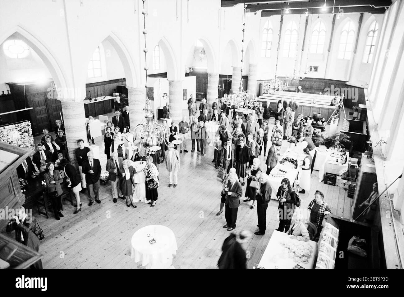 Eröffnung Bakenesserkerk Haarlem the Netherlands, Whizgle News, Dutch Desk, Niederlande, 1950 - 2000 auf 30-04-1997. Das Bild enthält diese Themen. Die Szene entfaltet sich in einem geräumigen Saal, der sich durch hohe, bogenförmige Decken und große Fenster auszeichnet, die weiches, diffuses Licht eindringen lassen und eine warme und einladende Atmosphäre schaffen. Die Wände, die mit Holztäfelungen verziert sind, verleihen der Umgebung einen Reichtum und stehen im Kontrast zu den hellen Tönen des Bodenbelags. Im Vordergrund befindet sich ein runder Tisch mit weißem Leinen, umgeben von einer Reihe von Gästen, die in halbformalen Kleidern gekleidet sind. Stockfoto