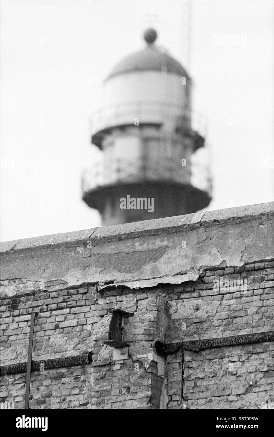 Diverse Hafengegend, Whizgle News, Dutch Desk, Niederlande, 1950 - 2000 am 29-03-1993. Das Bild enthält diese Themen. Die Szene zeigt eine verwitterte Ziegelwand im Vordergrund, die sich durch ihre raue Textur und sichtbare Risse auszeichnet, die auf Alter und Vernachlässigung hinweisen. Verschiedene Grautöne dominieren die Ziegeloberfläche, wobei einige Bereiche aufgrund von Schattenspielen dunkler erscheinen, was auf ein Gefühl von Verfall hindeutet. Hoch im Hintergrund steht ein Leuchtturm, dessen abgerundeter Turm mit einer markanten Kuppel gekrönt ist. Der Leuchtturm verfügt über ein Geländer, das eine Aussichtsplattform anzeigt, und hat ein Gebrauchsmuster Stockfoto