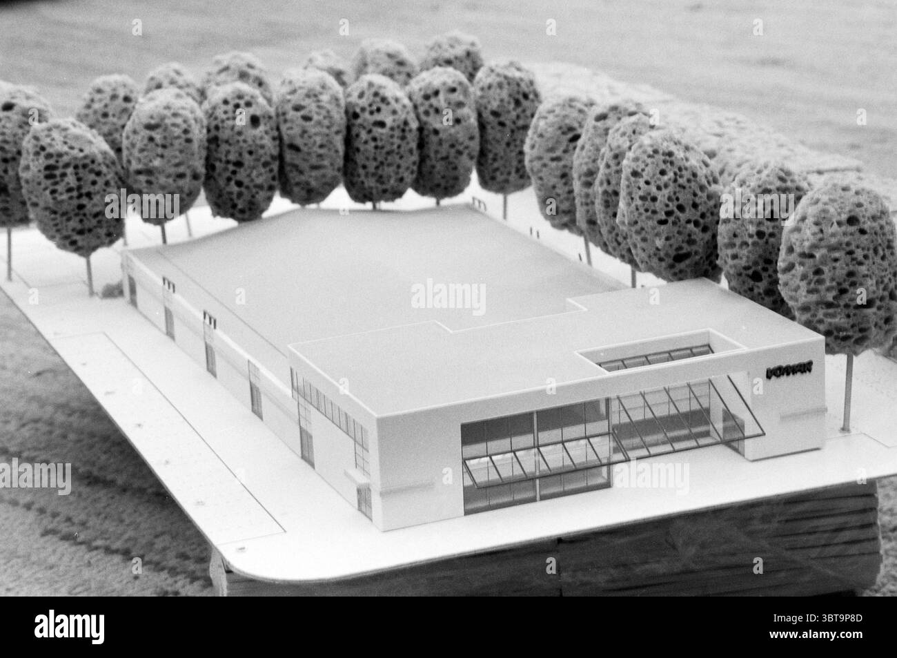 Modell für IJmuiden Modell IJmuiden Niederlande, Whizgle News, Dutch Desk, Niederlande, 1950 - 2000 am 08-12-1987. Diese Themen werden in der Abbildung angezeigt. Die Szene zeigt ein maßstabsgetreues Modell eines modernen Gebäudes, das in der Mitte deutlich positioniert ist. Die Struktur besticht durch klare Linien und ein kantiges Design, das einen minimalistischen architektonischen Stil betont. Seine Fassade ist überwiegend weiß mit großen Glasfenstern, die ein modernes Ambiente vermitteln und ein Gefühl von Offenheit schaffen. Um das Gebäude herum sind sorgfältig gearbeitete Modellbäume, die Schwämmen in der Textur ähneln. Diese Bäume mit ihren weichen, Stockfoto