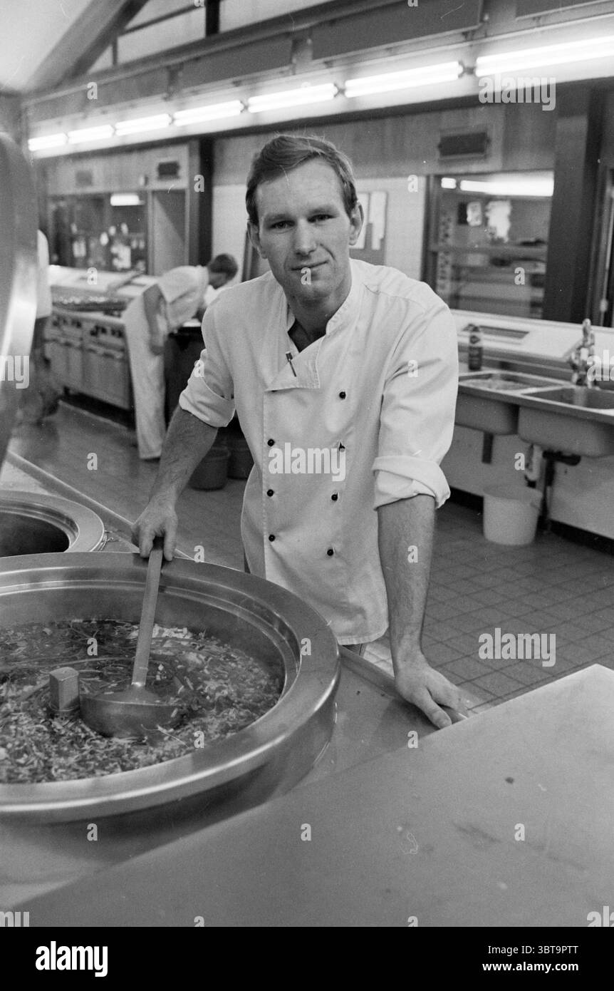 Herr Tol Cook Elizabeth Gasthuis Hospitals, Whizgle News, Dutch Desk, Niederlande, 1950 - 2000 am 09-12-1982. Das Bild enthält diese Themen. In einer belebten Küche steht ein Mann selbstbewusst neben einem großen Metalltopf und rührt eine dicke Mischung mit einer Holzkelle um. Er trägt eine klassische Küchenuniform mit einem zweireihigen weißen Mantel, der einen starken Kontrast zu den glänzenden Stahloberflächen um ihn herum bildet. Sein Ausdruck ist fokussiert und leicht stolz, was auf ein Gefühl der Leistung in seinen kulinarischen Bemühungen hindeutet. Die Küche ist gut beleuchtet mit hellen Deckenleuchten, die ein warmes gl erzeugen Stockfoto