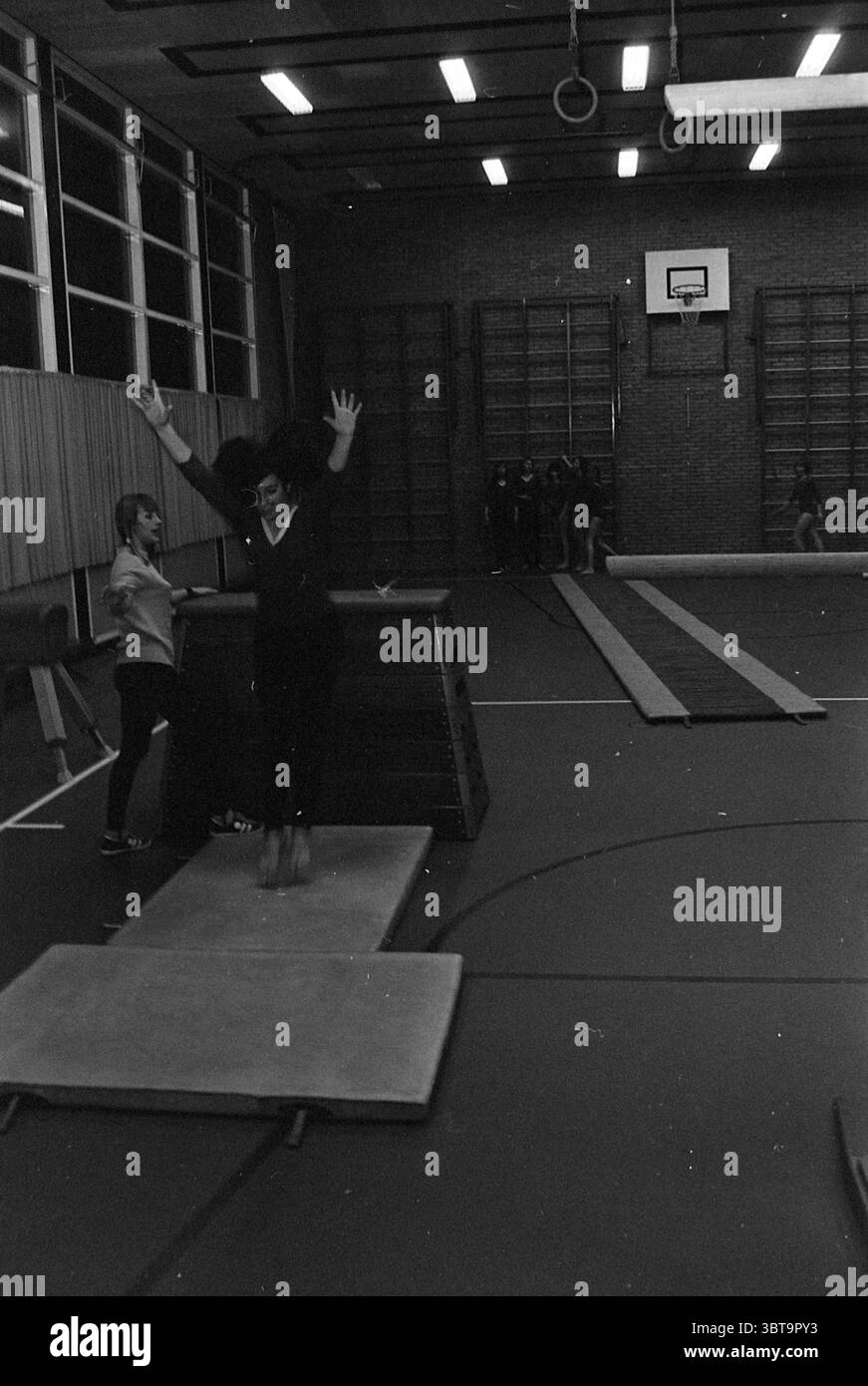 Gymnastikverband IJmuiden Gymnastics, Whizgle News, Dutch Desk, Niederlande, 1950 - 2000 am 15-02-1972. Das Bild enthält diese Themen. In einem geräumigen Fitnessraum ist die Szene mit einer Atmosphäre voller Konzentration und Aktivität erfüllt. Die Wände sind mit Holzvertäfelungen verziert, und die hohen Fenster lassen ein gedämpftes Licht in den braunen Boden des Fitnessstudios strahlen. In der Mitte wird eine Frau mitten in der Leistung auf einem Balancebalken erfasst, ihre Arme sind hoch angehoben, um das Gleichgewicht zu erreichen, was ein Gefühl von Entschlossenheit und Anmut vermittelt. Sie trägt ein dunkles Outfit, das sich mit den helleren Farben des Fitnessstudios abhebt. Ihr Ausdruck Stockfoto