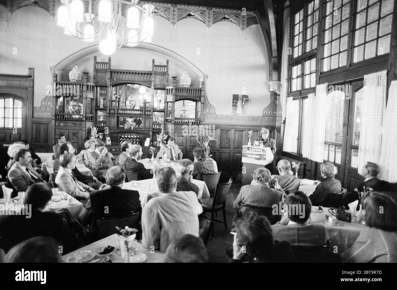 Eröffnung der Station Haarlem Eröffnung der Öffnungen Inbetriebnahme Station Haarlem Niederlande, Whizgle News, Dutch Desk, Niederlande, 1950 - 2000 am 11-05-1989. Diese Themen sind in der Abbildung dargestellt. Die Szene entfaltet sich in einem elegant dekorierten Speisesaal, der von hohen Holzbalken und kunstvollen Details geprägt ist, die einen Sinn für historischen Charme widerspiegeln. Die Wände sind mit komplizierten Mustern verziert, während große Fenster natürliches Licht eindringen lassen und trotz der monochromatischen Töne der Umgebung ein weiches, warmes Ambiente schaffen. Im Vordergrund steht ein Sprecher auf einem einfachen Podium und spricht an Stockfoto