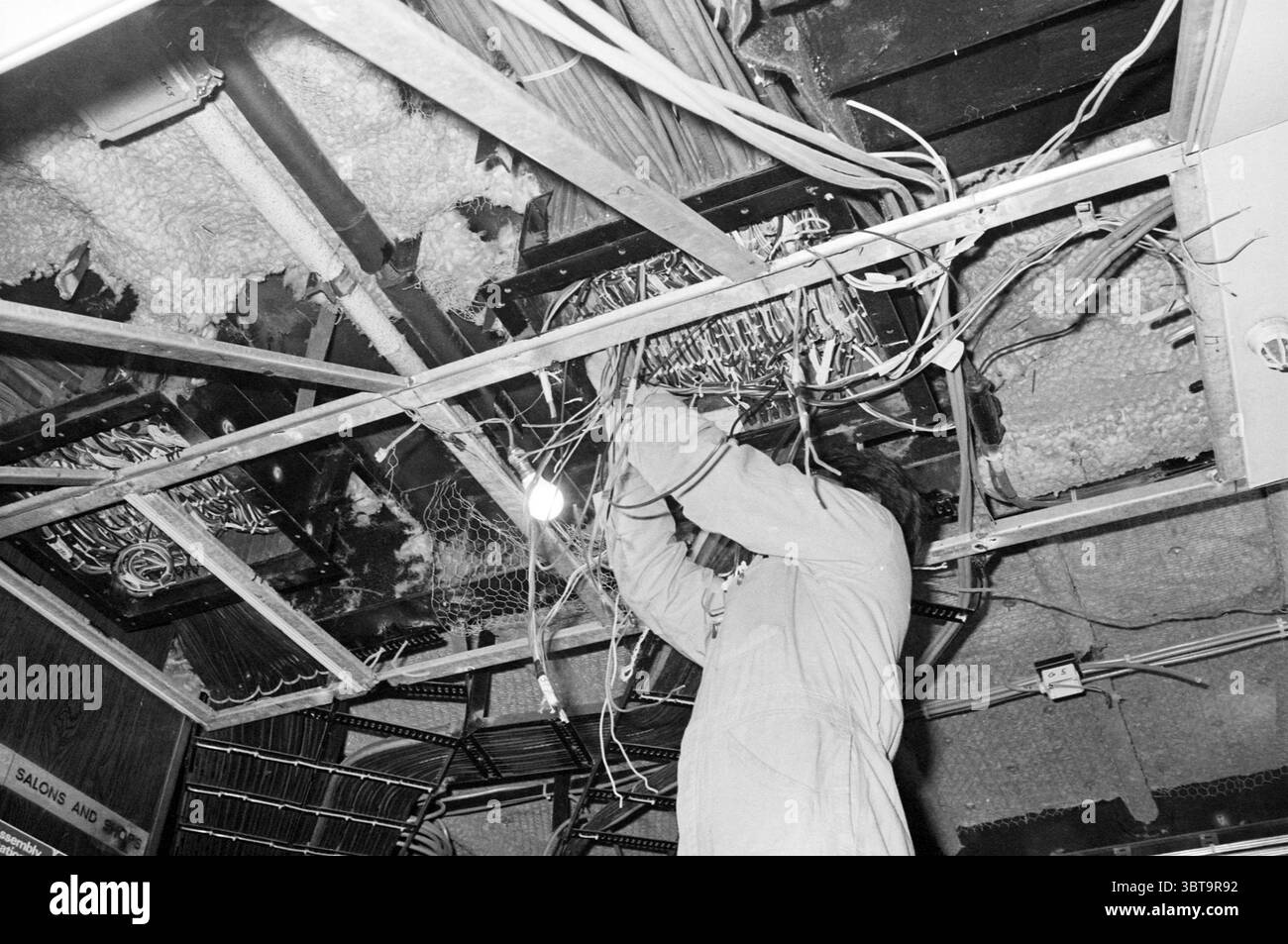 A.D.M. Schiffsbau Amsterdam Niederlande, Whizgle News, Dutch Desk, Niederlande, 1950 - 2000 am 15-01-1982. Das Bild enthält diese Themen. In einer schwach beleuchteten Umgebung wird eine Person, die in einem hellen Schutzanzug gekleidet ist, über ihnen eine Aufgabe ausgeführt, die bis in einen zerlegten Bereich reicht. Die Szene zeichnet sich durch ein chaotisches Gewirr von Drähten und Kabeln aus, die locker hängen und etwas ausgefranst wirken. Die Strukturen der Materialien – Metall, Kunststoff und möglicherweise Isolierung – bilden einen starken Kontrast zum glatten Material des Anzugs. Der Hintergrund wird von dar dominiert Stockfoto