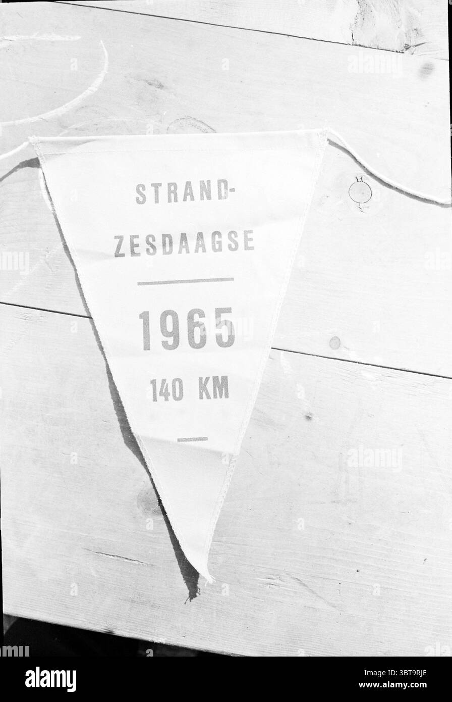 Six Day Beach Walking, Whizgle News, Dutch Desk, Niederlande, 1950 - 2000 am 04-08-1965. Das Bild enthält diese Themen. Die Szene zeigt eine dreieckige weiße Stofffahne, die ein leicht abgenutztes Aussehen hat und auf ihr Alter und ihre Verwendung hinweist. Der Stoff hat eine dezente Textur, die darauf hindeutet, dass er aus Baumwolle oder einem ähnlichen Material besteht, und er ist über eine helle Holzoberfläche, möglicherweise einen Tisch oder ein Brett, gewickelt. Die Flagge enthält einige gedruckte Details: Zahlen, die ein Jahr und eine Distanz anzeigen, die in einer fetten, unauffälligen Schrift dargestellt werden, die sich deutlich vom hellen Hintergrund abhebt. Der Text wird angezeigt Stockfoto