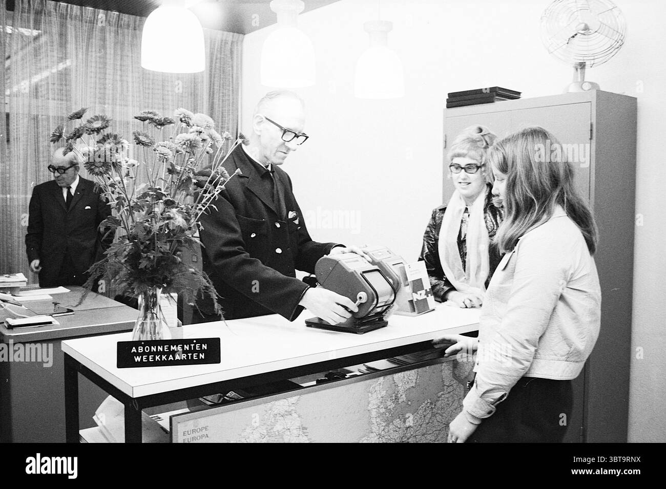 Cebuto NZH Noord Zuid Hollandse Vervoersmaatschappij N.V. NZ, Whizgle News, Dutch Desk, Niederlande, 1950 - 2000 am 28-11-1970. Diese Themen sind in der Abbildung dargestellt. In einem Retro-Setting ist ein dedizierter Zähler der Mittelpunkt, bei dem ein Mann in dunkler Uniform eine Aufgabe mit einer altmodischen Maschine ausführt. Seine Brille und sein gekämmtes Haar deuten auf Autorität und Professionalität hin. Die Theke selbst ist einfach und dennoch effektiv und verfügt über eine klare weiße Oberfläche vor einem Hintergrund gedämpfter Farben. Rechts eine junge Frau mit langen Haaren und einer leichten, figurbetonten Jacke Stockfoto