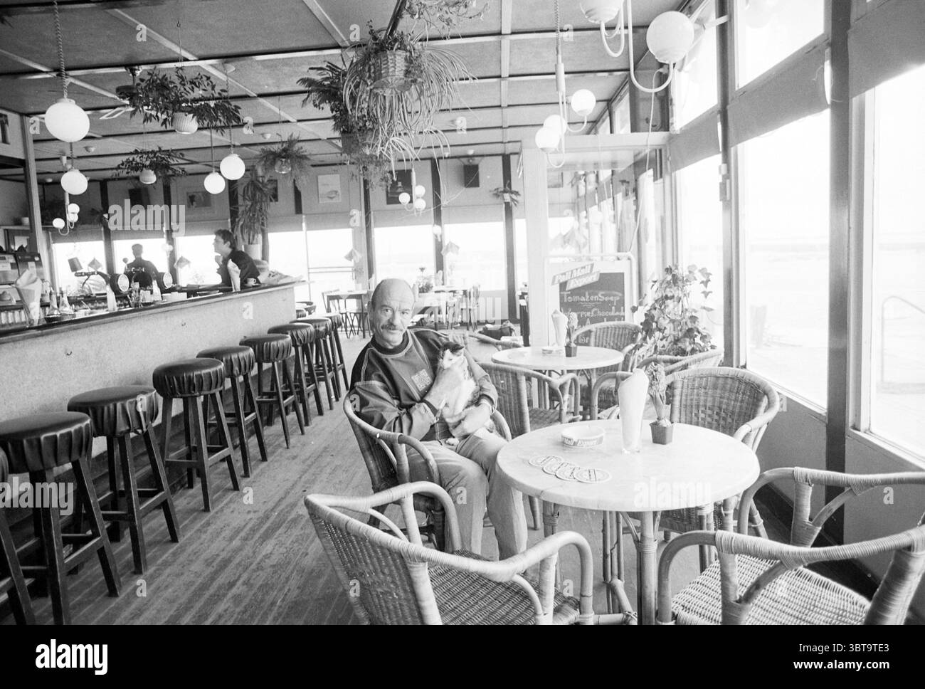 Strandpavillon 't Schouwtje Meindert, Whizgle News, Dutch Desk, Niederlande, 1950 - 2000 am 28-01-1992. Dies sind die Elemente im Bild. Die Szene fängt ein gemütliches, minimalistisches Café-Ambiente ein, das sich durch eine helle und luftige Atmosphäre auszeichnet. Die großzügigen Fenster lassen weiches, diffuses Sonnenlicht eindringen und beleuchten den Innenraum mit einem sanften, warmen Licht. Der Boden hat eine rustikale Holzoberfläche, die das einladende Ambiente unterstreicht. In der Mitte sitzt ein Mann bequem in einem gewebten Stuhl, seine entspannte Haltung deutet auf Freizeit hin. Er wiegt einen kleinen Hund in seinen Armen, ihre Bindung schafft einen herzerwärmenden Fo Stockfoto