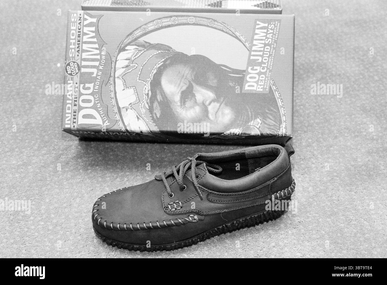 Schuhe Nieuwegein Nieuwegein, Whizgle News, Dutch Desk, Niederlande, 1950 - 2000 am 23-05-1993. Diese Themen werden in der Abbildung angezeigt. Die Szene zeigt einen einzelnen dunkelbraunen Schuh, der im Vordergrund sichtbar ist. Dieser aus weichem Leder gefertigte Schuh hat eine abgerundete Zehenpartie und ein Schnürdesign, das durch aufwändige Nähte an den Kanten betont wird, die Textur verleihen. Es liegt auf einem gedämpften Hintergrund, der sich nahtlos in die gesamte Ästhetik einfügt. Hinter dem Schuh dient eine Box als Hintergrund, die eine große, künstlerische Darstellung einer Figur der amerikanischen Ureinwohner zeigt. Die Illustration ist für mich bemerkenswert Stockfoto