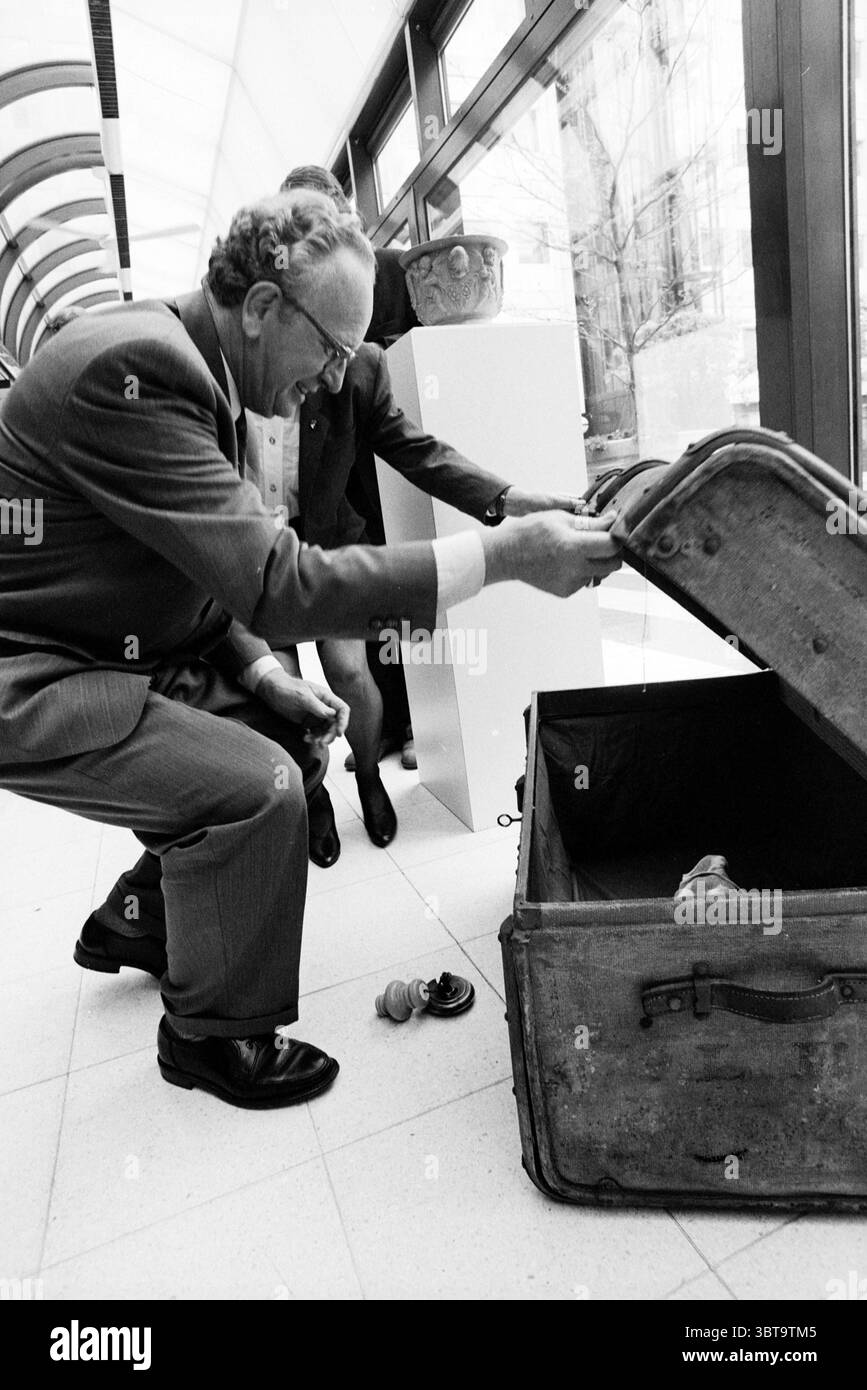 Eröffnungsausstellung K V K, Whizgle News, Dutch Desk, Niederlande, 1950 - 2000 am 30-06-1992. Diese Themen werden in der Abbildung angezeigt. Die Szene fängt einen Moment in einem modernen Innenraum ein, der sich durch klare Linien und eine helle, luftige Atmosphäre auszeichnet. Natürliches Licht strömt durch große Fenster hinein und wirft weiche Schatten über den Boden, der aus hellen Fliesen besteht. Im Vordergrund lehnt sich ein älterer Mann, in einem formellen Anzug gekleidet, aufmerksam nach vorne. Der Anzug ist ein klassisches Grau mit einem Hauch von Glanz. Sein Haar ist leicht zerzaust, was auf eine Mischung aus Fokus und Aufregung hindeutet, während er sich neben einer Vinta hockt Stockfoto