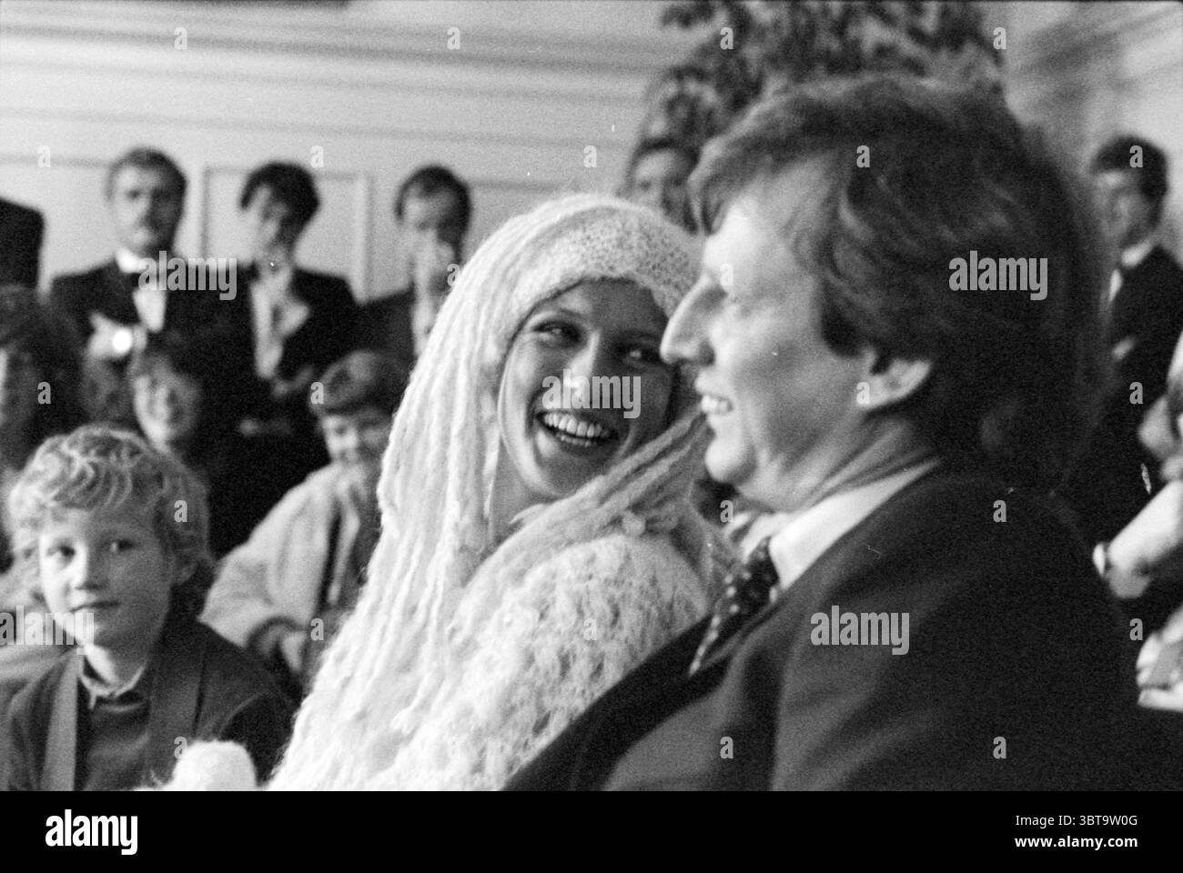 Marriage Udo kauft Marriage, Whizgle News, Dutch Desk, Niederlande, 1950 - 2000 am 24-11-1979. Diese Themen werden in der Abbildung angezeigt. Die Szene entfaltet sich in einer intimen Innenumgebung, gefüllt mit einer Zusammenkunft von Menschen, die alle in formeller Kleidung gekleidet sind, was auf einen besonderen Anlass hinweist. Die Beleuchtung ist weich und warm und sorgt für eine gemütliche Atmosphäre mit dezenten Schatten, die dem Raum Tiefe verleihen. An der Spitze wird eine Frau in einem Moment der Freude gefangen gehalten, ihr weites Lächeln beleuchtet ihr Gesicht. Sie trägt ein einzigartiges, strukturiertes Outfit – möglicherweise ein gestricktes Kleidungsstück –, das elegant drapiert und den verspielten Charakter ihrer E unterstreicht Stockfoto