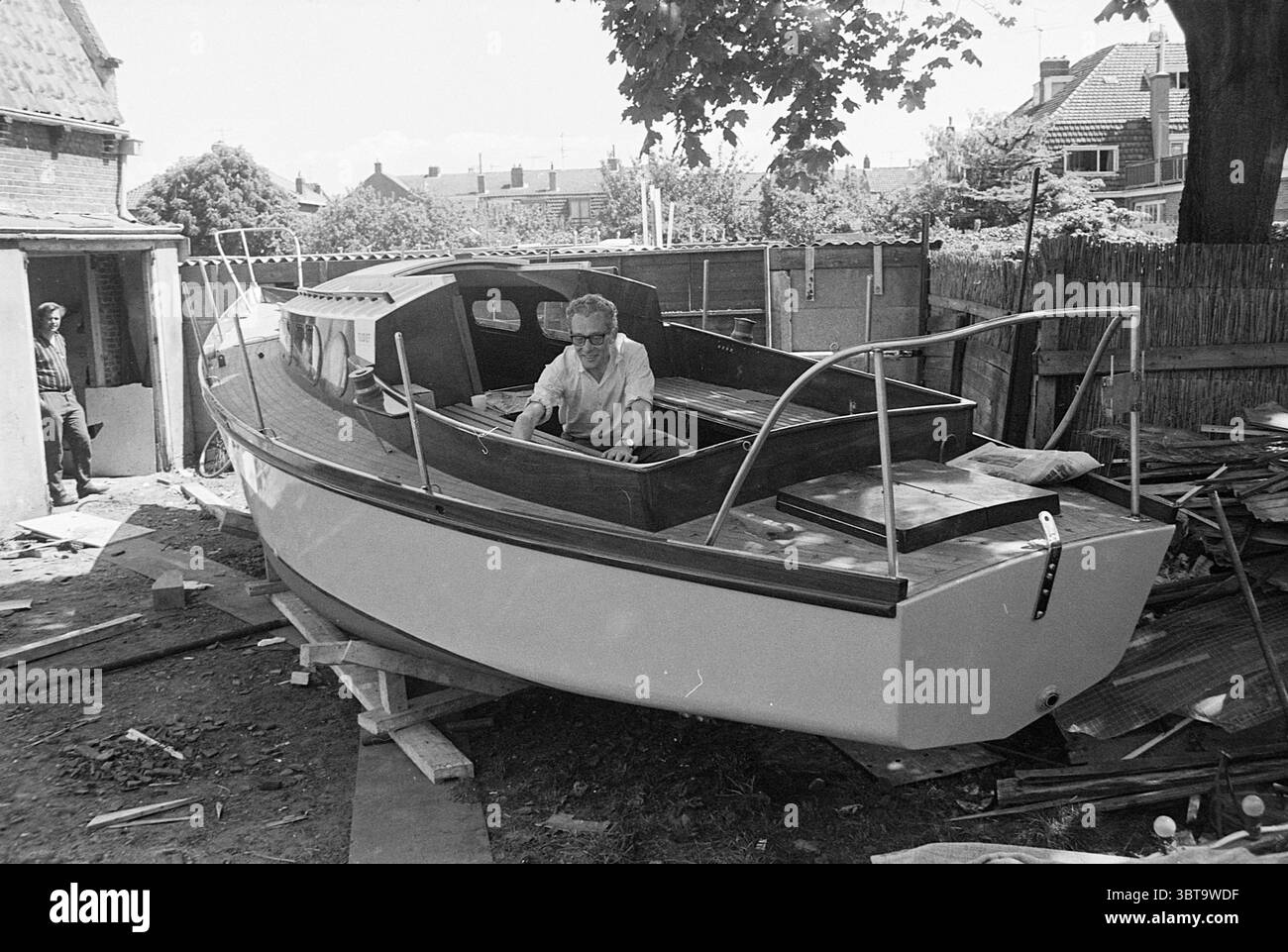 Herr van Gasteren bei Boat Persons, Whizgle News, Dutch Desk, Niederlande, 1950 - 2000 am 29.05.1969. Diese Themen sind in der Abbildung dargestellt. Die Szene fängt einen Moment in einer Werft ein, in dem sich Handwerkskunst und harte Arbeit kreuzen. In der Mitte nimmt ein schlankes, teilweise fertiggestelltes Boot Form an, dessen Rumpf in einem sauberen weiß lackiert ist, das sich von den Erdtönen der Umgebung abhebt. Das Boot ist auf Holzstützen angehoben, was auf laufende Bau- und Wartungsarbeiten hindeutet. Ein Mann sitzt am Steuer, konzentriert und engagiert in seine Aufgabe. Er trägt eine Brille, und sein Ausdruck zeigt Konzentrik Stockfoto
