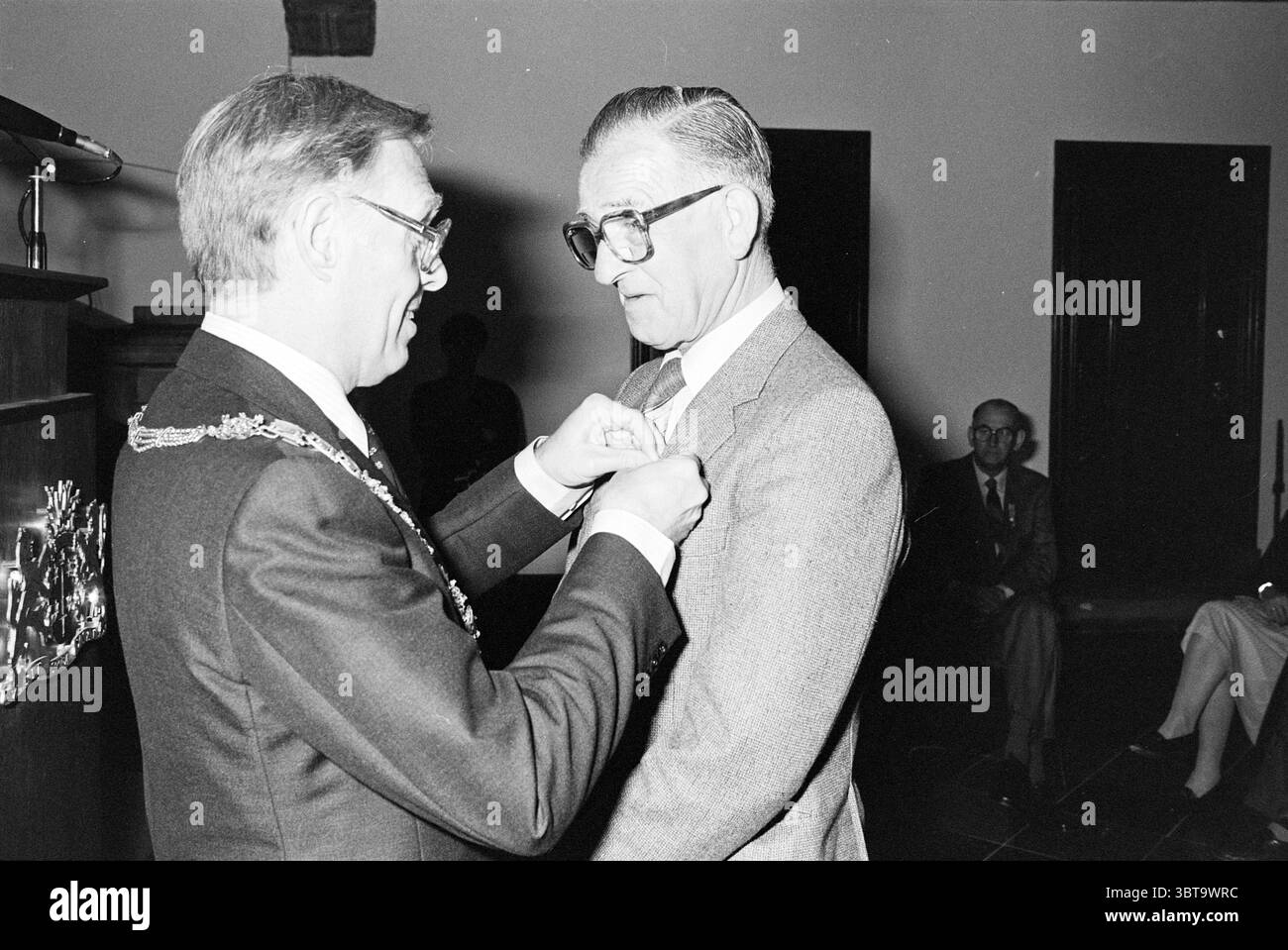 Award Municipality of Haarlem Awards Haarlem Grote Markt Niederlande, Whizgle News, Dutch Desk, Niederlande, 1950 - 2000 am 29-04-1982. Dies sind die Themen im Bild. Die Szene fängt ein formelles Ereignis ein, das in Innenräumen stattfindet und von einem bemerkenswerten Hauch von Professionalität und Feierlichkeiten geprägt ist. Zwei Personen stehen im Mittelpunkt: Einer ist mit Anzug und Brille verziert, der Akt, ein Abzeichen oder eine Medaille auf das Revers des anderen zu stecken, der ebenfalls in einen formellen Anzug gekleidet ist und ein Bild der Ehre und Anerkennung projiziert. Die Einrichtung verfügt über ein elegantes Interieur mit dunkler Holzscheibe Stockfoto