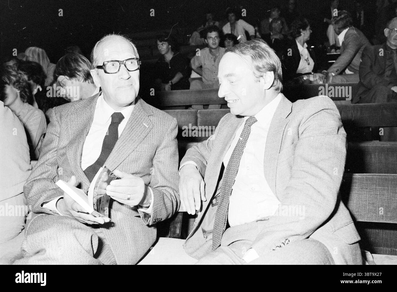 Filmbuchpräsentation in 't Hoogt Presentation Spectrum Utrecht, Whizgle News, Dutch Desk, Niederlande, 1950 - 2000 am 28-09-1981. Das Bild enthält diese Themen. In einer schwach beleuchteten Umgebung sitzen zwei Männer eng zusammen auf Holzbänken und unterhalten sich. Die Atmosphäre wirkt warm und intim, was auf einen gemeinsamen Moment der Kameradschaft hindeutet. Der Mann auf der linken Seite, der eine runde Brille trägt, hat einen nachdenklichen Ausdruck, begleitet von einem ordentlich gestylten Glatzkopf und einem hellgrauen Anzug mit dezenter Textur. In seinen Händen hält er ein kleines, geschlossenes Buch, das auf eine wissenschaftliche oder reflektierende Natur hinweist. Stockfoto