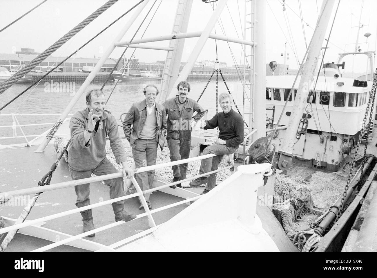 Fischerboot IJmuiden115 mit Besatzung IJmuiden Harbour Fisheries und andere Fischhändler Fischen IJmuiden Niederlande, Whizgle News, Dutch Desk, Niederlande, 1950 - 2000 am 06-05-1985. Das Bild enthält diese Themen. Die Szene zeigt eine Gruppe von vier Individuen, die auf dem Deck eines Fischereifahrzeugs stehen. Sie tragen praktische Arbeitskleidung – schwere Jacken, Overalls und Stiefel –, die auf ihr Engagement in der maritimen Arbeit hinweist. Ihre Ausdrücke vermitteln eine Mischung aus Kameradschaft und Stolz, was auf einen Moment der Verwandtschaft in einem harten Tag der Arbeit hindeutet. Der Hintergrund zeigt den Rumpf des Bootes, Farbe Stockfoto