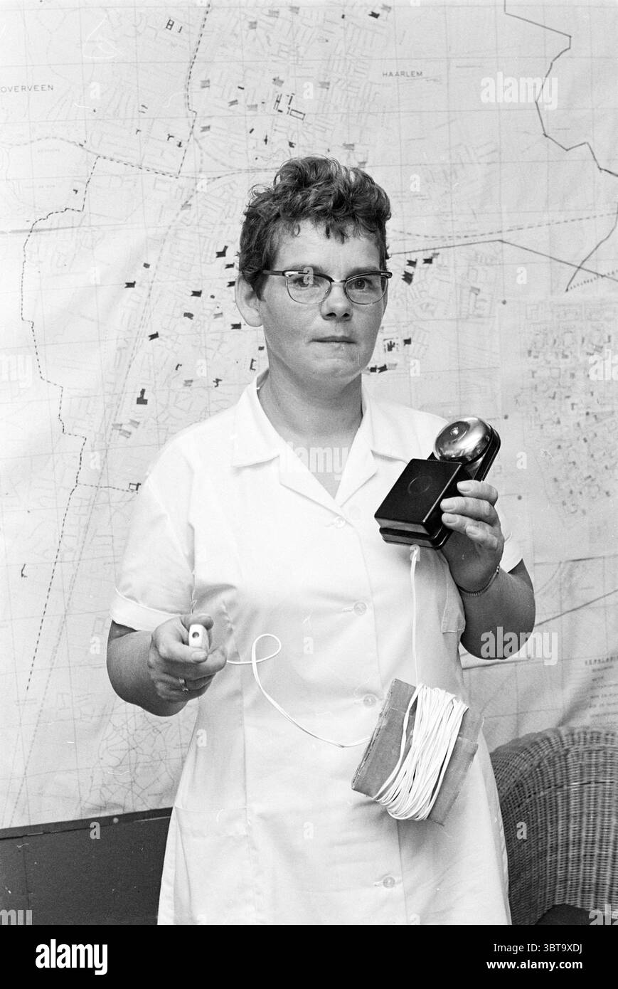 Frau mit Glocke in weißer Uniform., Whizgle News, Dutch Desk, Niederlande, 1950 - 2000. Das Bild enthält diese Themen. In der Szene steht eine Frau im Vordergrund, die eine alte Kamera in der einen Hand und ein kleines Gerät in der anderen hält. Sie hat kurze, ordentlich gestylte Haare und trägt eine runde Brille, was ihr ein gelehrtes Aussehen verleiht. Ihr Outfit besteht aus einem hellen, kurzärmeligen Kleid, das typisch für die Mitte des 20. Jahrhunderts ist. Die Kulisse zeigt eine große, detaillierte Karte, die wahrscheinlich ein Stadtbild darstellt, mit zahlreichen Linien, Markierungen und geografischen Merkmalen, die auf ein städtisches en hindeuten Stockfoto