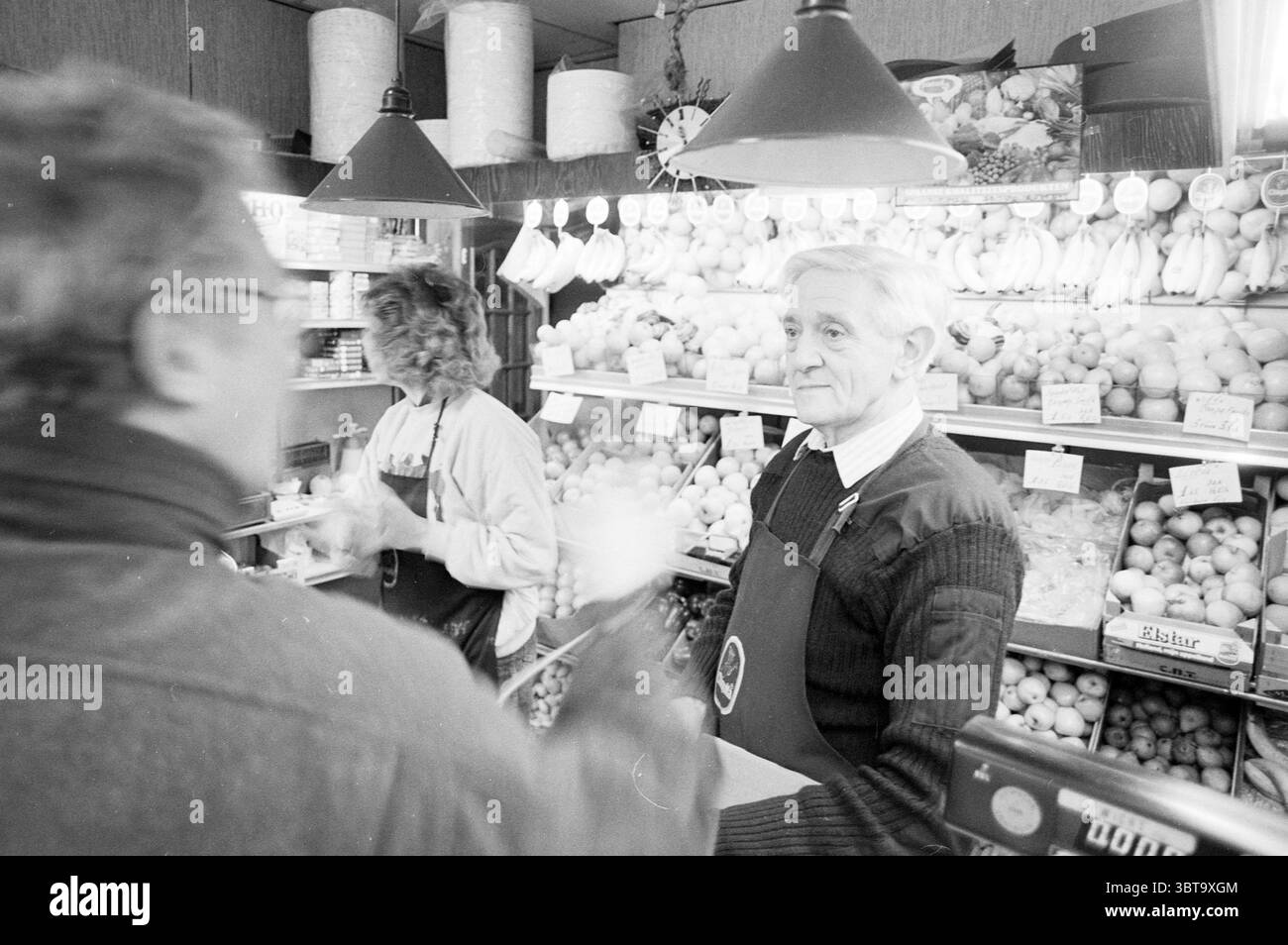 Interview mit Jaap Tanis Haarlem Smedestraat Niederlande, Whizgle News, Dutch Desk, Niederlande, 1950 - 2000 am 18.03.1989. Diese Themen werden in der Abbildung angezeigt. In einer belebten Marktszene führen zwei Personen eine lebhafte Interaktion an einem Obststand. Die Kulisse bietet eine lebhafte Präsentation frischer Produkte mit einer Auswahl an bunten Früchten wie Orangen, Bananen und Äpfeln, die ordentlich übereinander liegen. Die satten Farbtöne der Früchte stehen im Kontrast zu den gedämpften Tönen der Umgebung. Eine Person, ein älterer Gentleman, steht selbstbewusst hinter der Theke und trägt ein dunkles Schwitzen Stockfoto