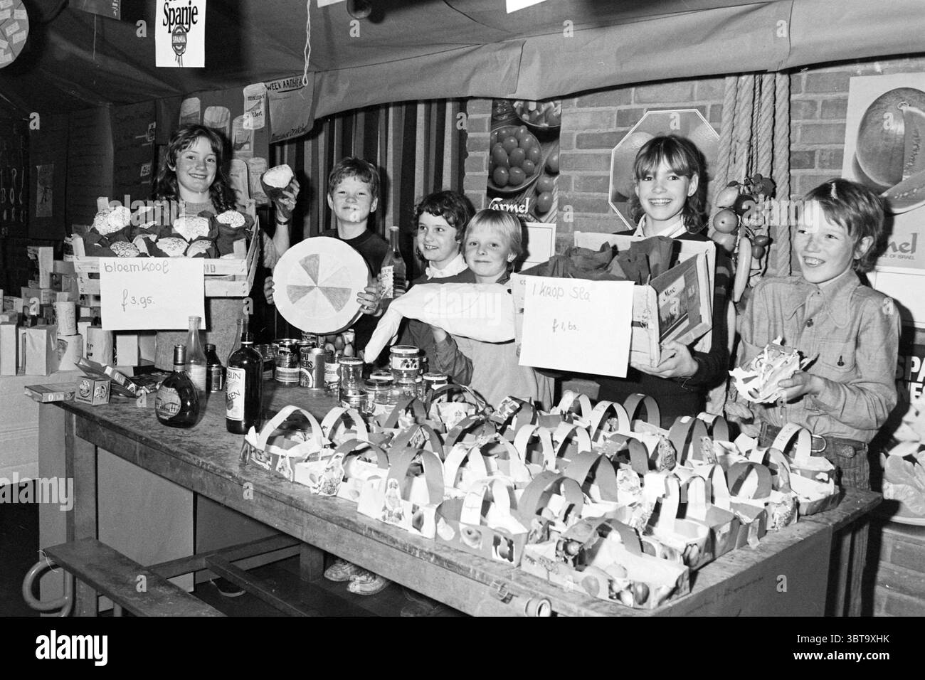 Ernährungsprojekt Pieter Vermeulen School IJmuiden Schools IJmuiden the Netherlands, Whizgle News, Dutch Desk, Niederlande, 1950 - 2000 am 29-01-1981. Das Bild enthält diese Themen. Die Szene fängt einen pulsierenden Marktstand voller Energie und jugendlicher Spannung ein. Hinter einem langen Tisch mit verschiedenen Waren steht stolz eine Gruppe von sechs Kindern, von denen jedes ein oder zwei Gegenstände zeigt, die den Charme der Anlage insgesamt noch verstärken. Der Stand ist mit einer Vielzahl von Produkten geschmückt, darunter buntes Obst und verpackte Waren. Der Tisch selbst ist eine rustikale Holzoberfläche, die einen warmen Stockfoto