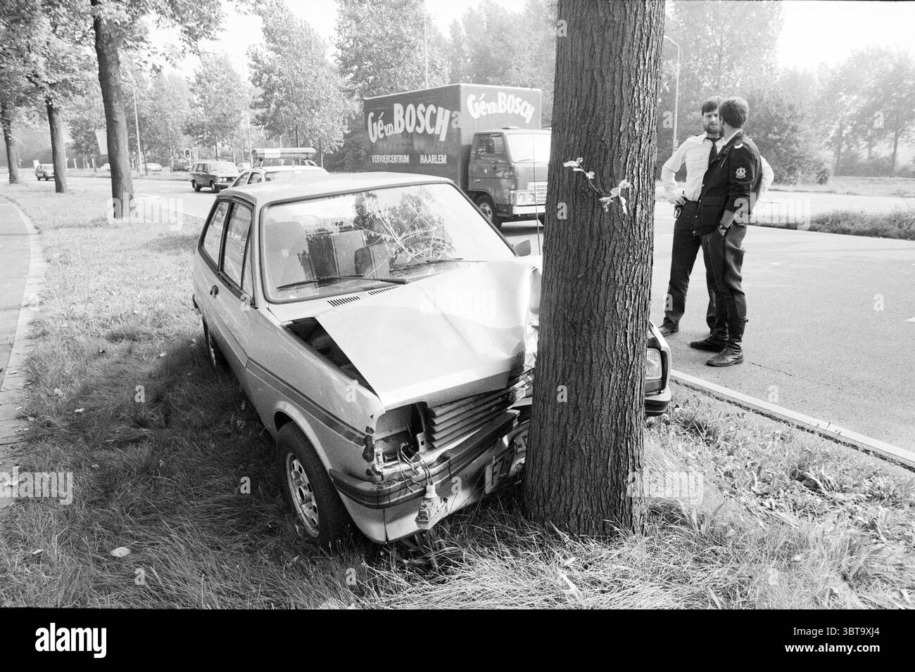 Verkehrsunfallwagen gegen Baum, Whizgle News, Dutch Desk, Niederlande, 1950 - 2000 am 09-1987. Dies sind die Elemente im Bild. Die Szene fängt einen düsteren Moment auf einer urbanen Straße ein. Ein kleines, helles Auto ist sichtbar beschädigt, seine Vorderseite ist an der Basis eines großen Baumes zerknittert, was auf einen Unfall vor kurzem hindeutet. Die Motorhaube des Fahrzeugs ist nach oben gebogen, und der vordere Stoßfänger ist teilweise abgenommen, wodurch sich Metall und Kunststoff vermischen. Um das Fahrzeug herum ist das Gras leicht bewachsen und mit Wildblumen gespickt, was einem ansonsten heftigen Vorfall einen Hauch von Natur verleiht. Im bac Stockfoto