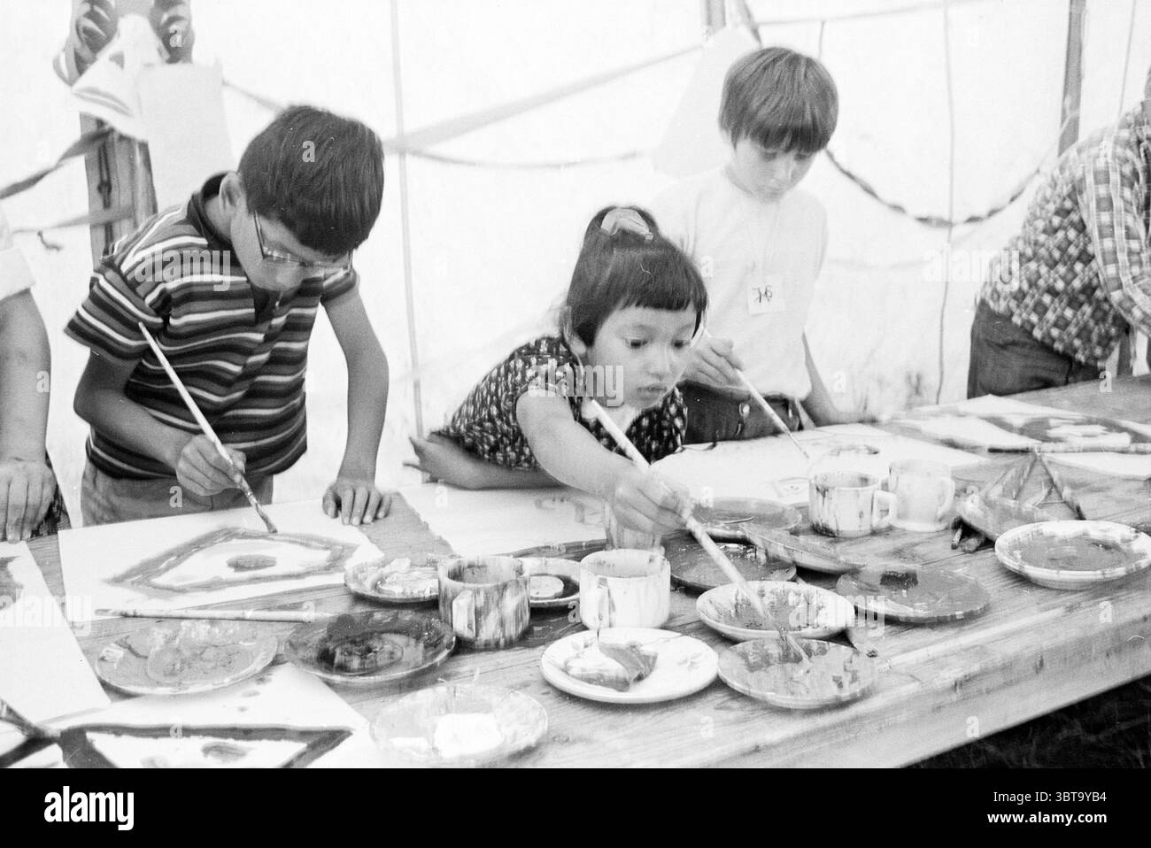 Färbung für Kinder Beverwijk Ferienspiele Ferientage Ferienlager, Whizgle News, Dutch Desk, Niederlande, 1950 - 2000 auf 09-07-1968. Die Abbildung zeigt diese Themen. Die Szene entfaltet sich in einem geräumigen, sanft beleuchteten Zelt und schafft eine warme und einladende Atmosphäre. Die Wände des Zeltes sind hell, sodass weiches Licht durchdringen kann, was das gesamte Ambiente verbessert. An der Vorderseite ist ein Holztisch mit großen Papierbögen und zahlreichen Farbpaletten bedeckt, die jeweils mit lebhaften Farben gefüllt sind. Der Tisch selbst ist leicht abgenutzt, was darauf hindeutet, dass er viele kreative Engagements erlebt hat Stockfoto