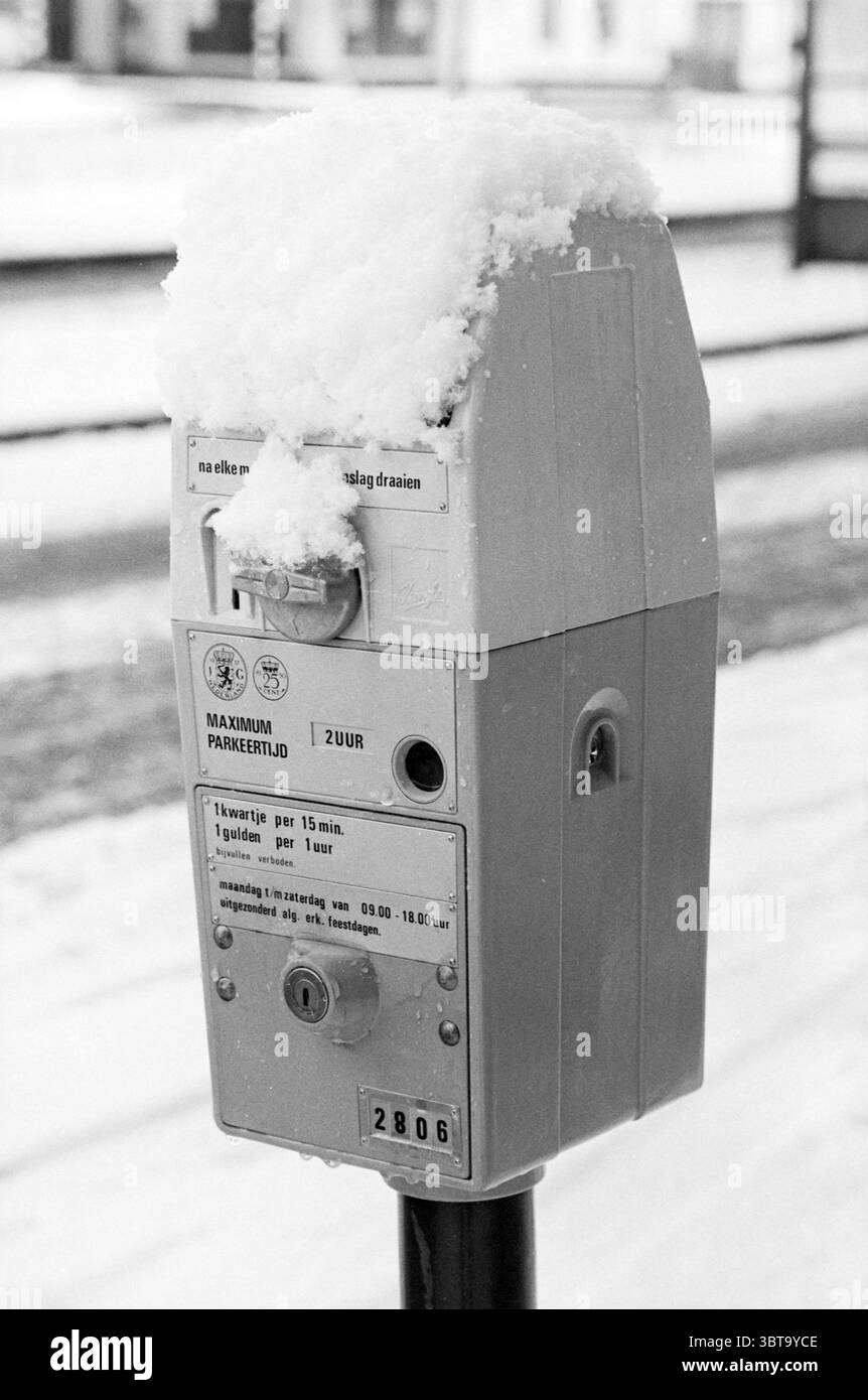 Snow Haarlem und Umgebung Snow Haarlem Niederlande, Whizgle News, Dutch Desk, Niederlande, 1950 - 2000 am 12-12-1981. Diese Themen werden in der Abbildung angezeigt. Im Vordergrund steht eine Parkuhr, deren Oberfläche teilweise mit einer frischen Schneeschicht bedeckt ist, was ihr eine weiche, weiße Kappe verleiht. Der Körper des Messgeräts ist ein gedämpftes Grau, das sich vom hellen weiß des Schnees abhebt. Die Umgebung scheint eine ruhige, urbane Straßenszene zu sein, wahrscheinlich bei kälterem Klima, wie die Winterbedingungen belegen. Der Hintergrund ist mit Weichzeichnungselementen verschwommen, die auf andere geparkte Fahrzeuge hinweisen Stockfoto